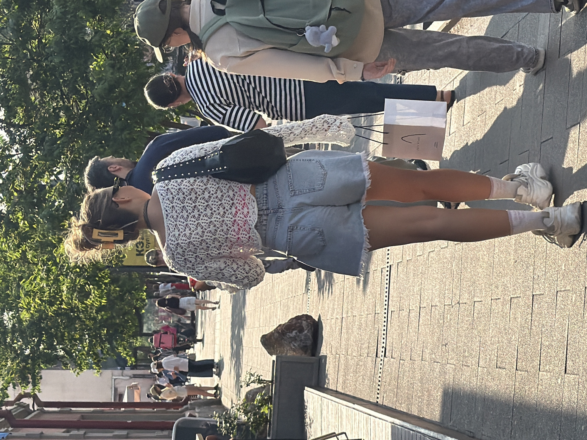 This is a street-level, medium shot of people walking on a sidewalk in Krasnoyarsk, Russia, likely during daylight hours as evidenced by the strong shadows. The focus is on a young woman in the foreground, seen from behind, wearing a white lacy crop top, denim shorts, white socks, and chunky white sneakers. She has her hair styled up with a large yellow clip. She is carrying a black handbag with silver stud details. To her right, a person wearing a striped shirt and dark pants walks behind her, carrying a paper shopping bag with brown and white sections. Further to the right, another person is visible from the back, wearing a light-colored jacket and a green backpack with a gray plush toy attached. In the background, several other individuals are walking in the same direction. Trees line the sidewalk, casting dappled shadows on the paved walkway. A large rock is placed near the left edge of the image, and planters are visible. The overall atmosphere appears to be casual and urban.