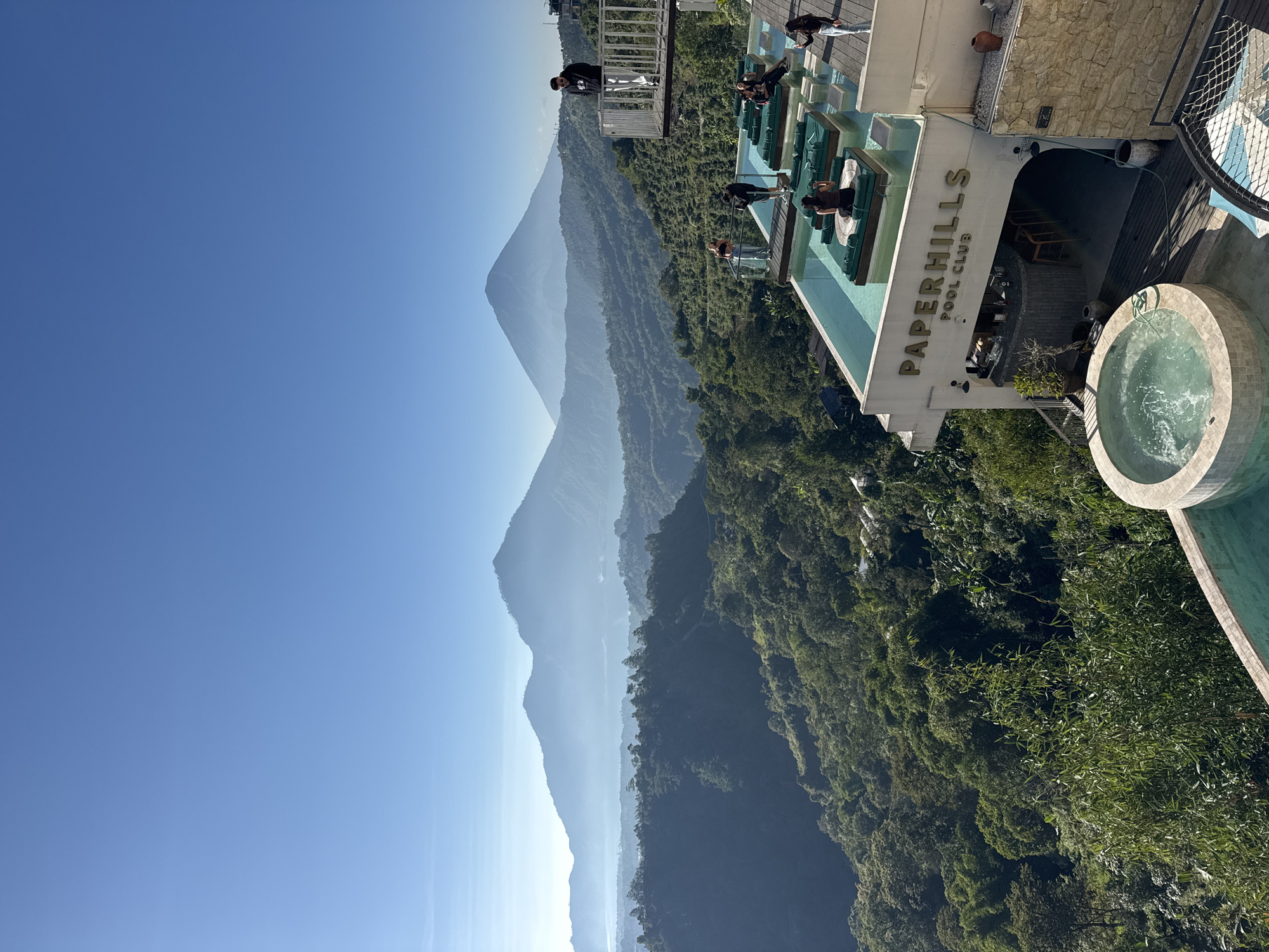 The image displays a panoramic outdoor scene from a high vantage point in Kintamani, Indonesia, under a clear blue sky. In the foreground, a multi-level establishment named "PAPERHILLS POOL CLUB" is visible. The building features light-colored concrete construction and a dark wooden deck. The lower level includes an arched entrance leading to a bar area with stools and bottles. Above this, the text "PAPERHILLS POOL CLUB" is affixed in gold lettering.
On the upper level of the building, a rectangular infinity pool contains several submerged or partially submerged dark green cushioned lounge platforms. At least four individuals are present in the pool, some seated or reclining on these platforms. To the right of the pool, on the wooden deck, one person in a black top and light pants is standing, holding a mobile device.
Below the main pool level, a circular, tiled hot tub with bubbling water is situated on the wooden deck. Further to the bottom right, a segment of a black net structure, possibly a trampoline or lounging net, is partially visible. Potted plants and decorative clay pots are placed on the various deck levels.
Above the main pool, a glass-paneled balcony extends, on which two individuals are standing, looking outwards. Higher up and to the right, a separate metal-railed balcony juts out from another part of the structure. One person in a black hoodie and light pants stands on this balcony, looking down or outwards.
The midground and background consist of a vast, mountainous, forested landscape. Numerous layers of green-covered mountains recede into the distance, exhibiting atmospheric haze that lightens their color. A large body of water, identifiable as Lake Batur, is situated amidst these mountains, partially obscured by mist or haze. Two prominent, conical peaks, characteristic of volcanoes in the Kintamani region, stand on the distant horizon against the clear blue sky. The sky is uniformly clear and light blue, indicating bright daylight conditions. In total, at least eight individuals are present across the various lounging and viewing areas.