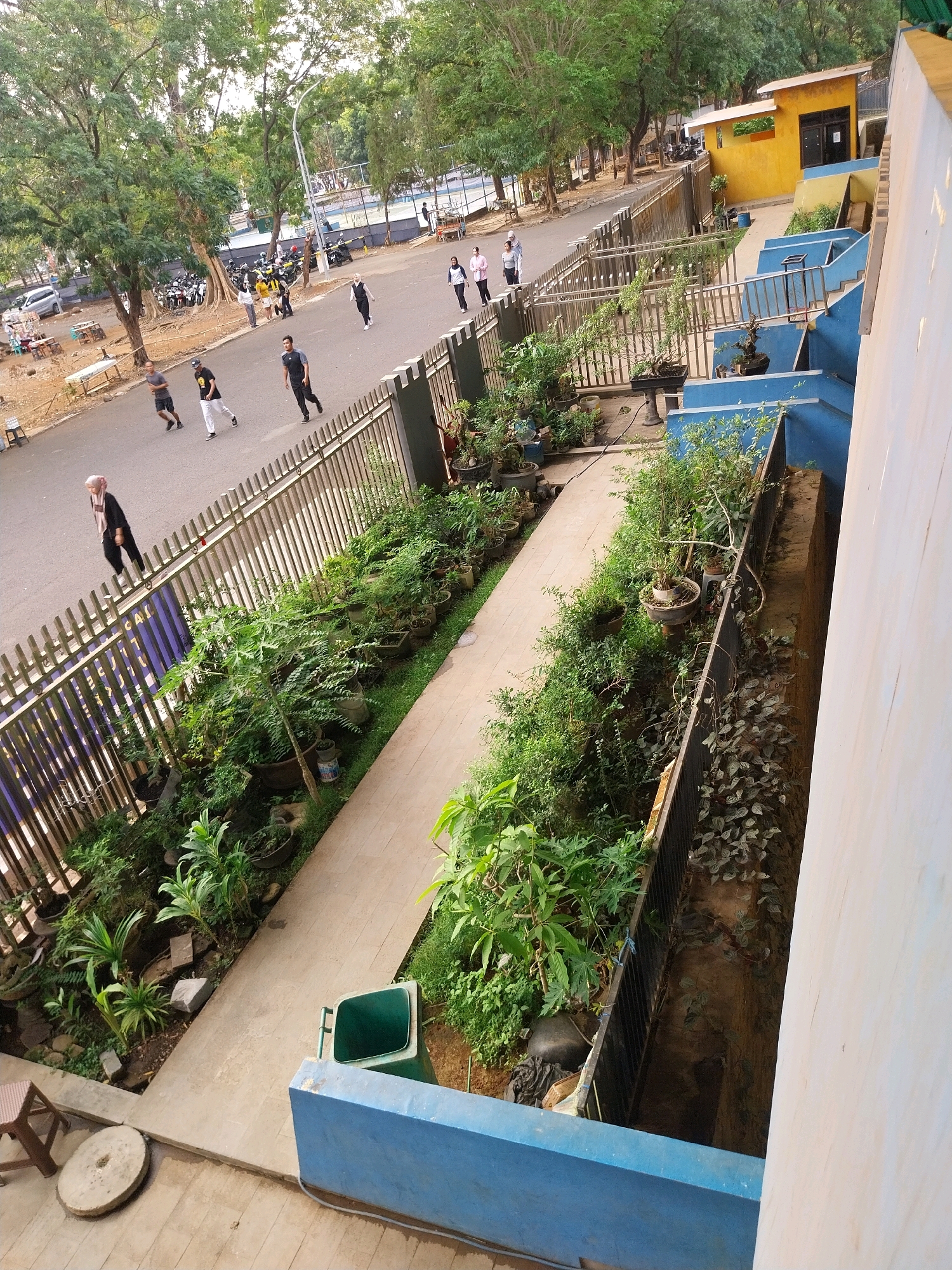 The image shows a street scene in Cirebon, Indonesia, with a paved road running alongside a fenced-off area with numerous potted plants and trees. Several people are visible walking and jogging on the sidewalk and road. In the background, there are parked motorcycles, a building with a yellow facade, and a tennis court. The overall impression is of an urban environment with green spaces incorporated into it.