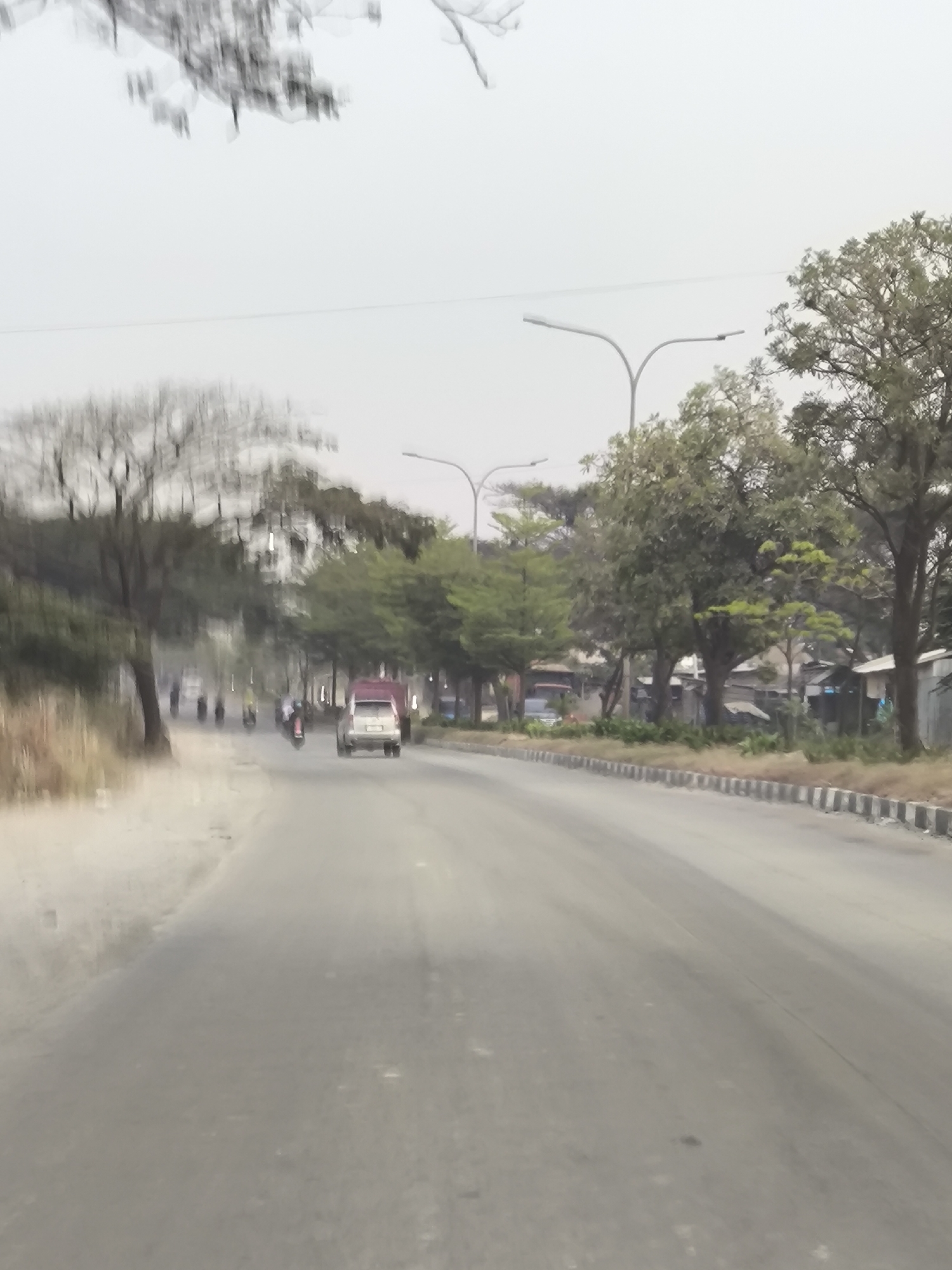 The image depicts a street scene in Cilegon, Indonesia, characterized by a cloudy sky and limited visibility due to a dusty atmosphere. A paved road dominates the foreground, stretching into the distance. On the right side of the road, a line of trees and some low-lying structures are visible. A single car, possibly a small SUV, is moving away from the camera in the right lane. On the left side of the road, a dry, sandy embankment is present, with a few sparse trees and brush. Further down the road, several motorcycles are traveling in the same direction as the car, with their occupants appearing as blurred figures. The overall impression is that of a moderately busy street on an overcast day.