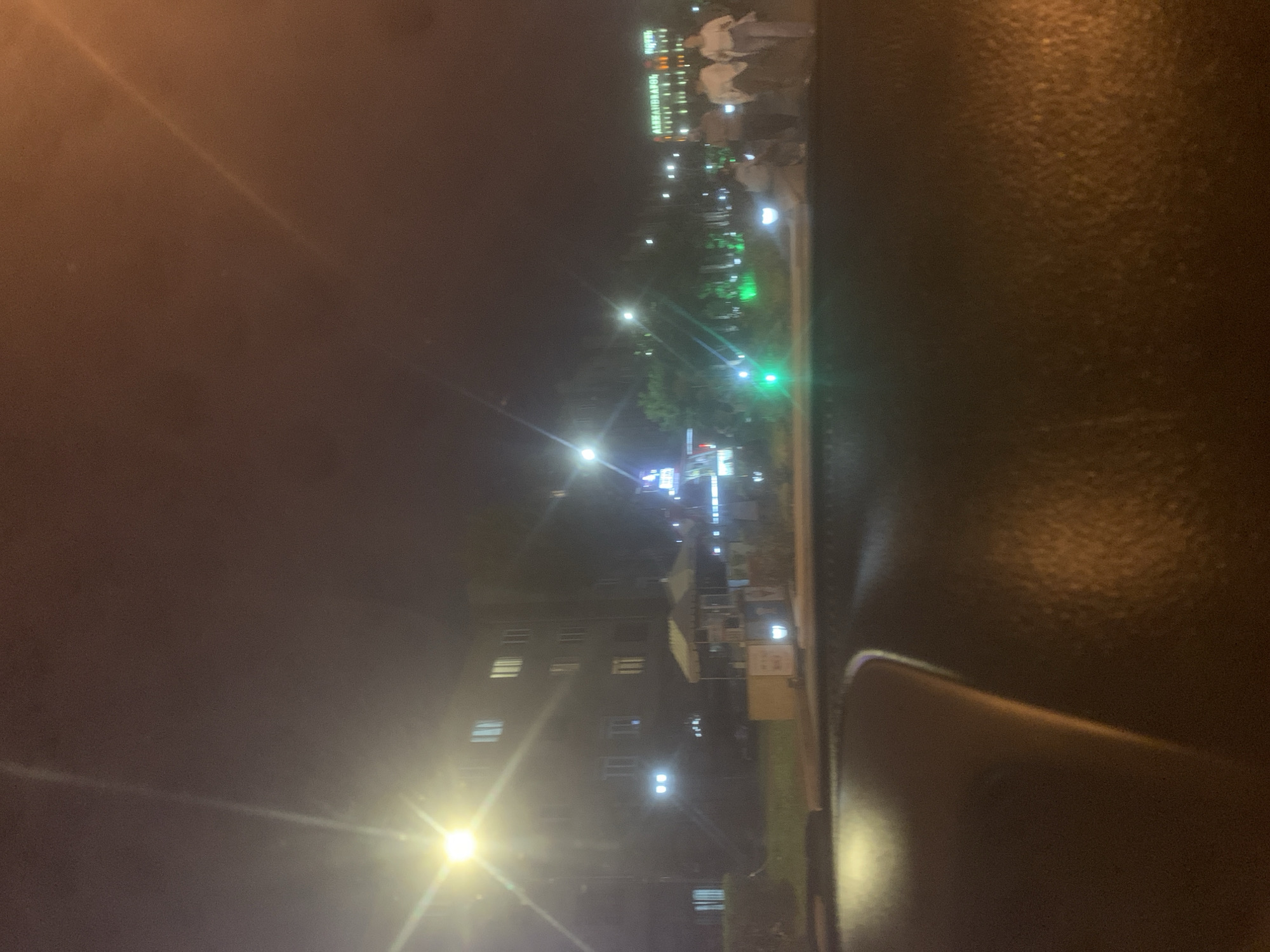 The image captures an urban night scene in Gyumri, Armenia, viewed from inside a vehicle through its windshield, evidenced by the reflective dashboard in the foreground. On the left, a multi-story building with multiple illuminated windows stands behind a bright streetlight, casting significant lens flare. Below the building, an outdoor area features two white canopied structures and several kiosks or stands. To the right, a street or open pedestrian area is visible, illuminated by various green ground lights and additional streetlights further down. In the background, a large building is identifiable as the "ALEXANDRAPOL HOTEL" through its illuminated signage. Several individuals, approximately four to five, are seen walking across the right side of the frame near the hotel, their forms blurred by distance and motion. The overall environment is an active city street at night, characterized by a mix of commercial and hospitality structures, artificial lighting, and pedestrian presence.