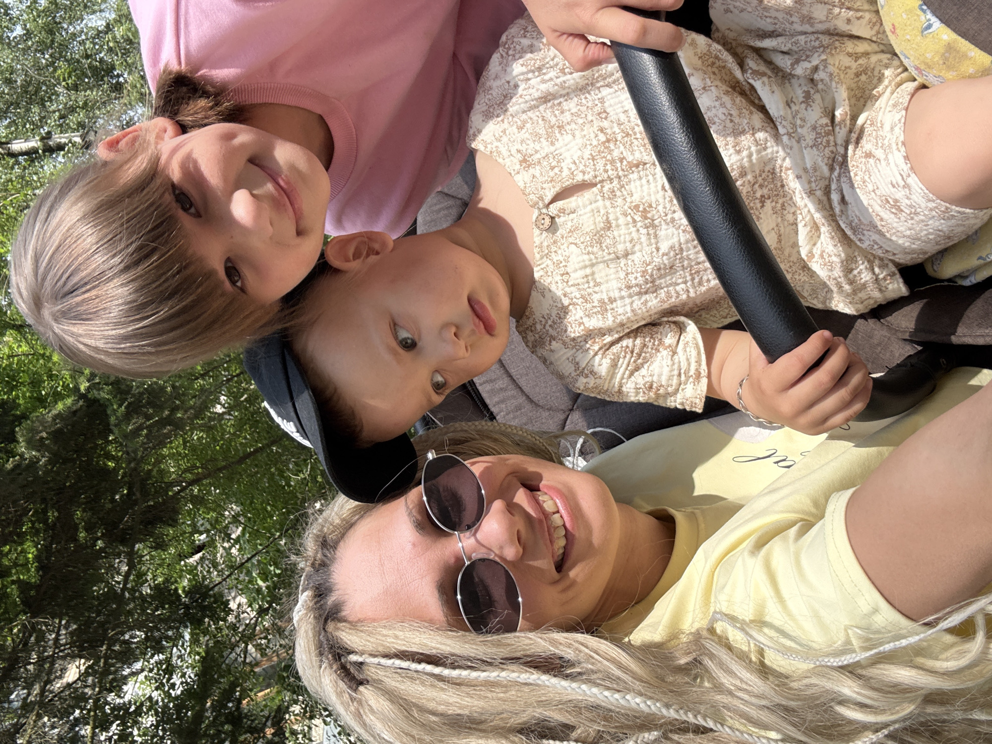 Three people are in the image. A woman with blonde braided hair and sunglasses, wearing a yellow shirt, is in the foreground. Above her and slightly to the left is a child wearing a pink shirt. To the right of the child is another child wearing a light-colored, patterned shirt and holding onto a black bar. Trees are visible in the background. The location is Шебунино, Russia.