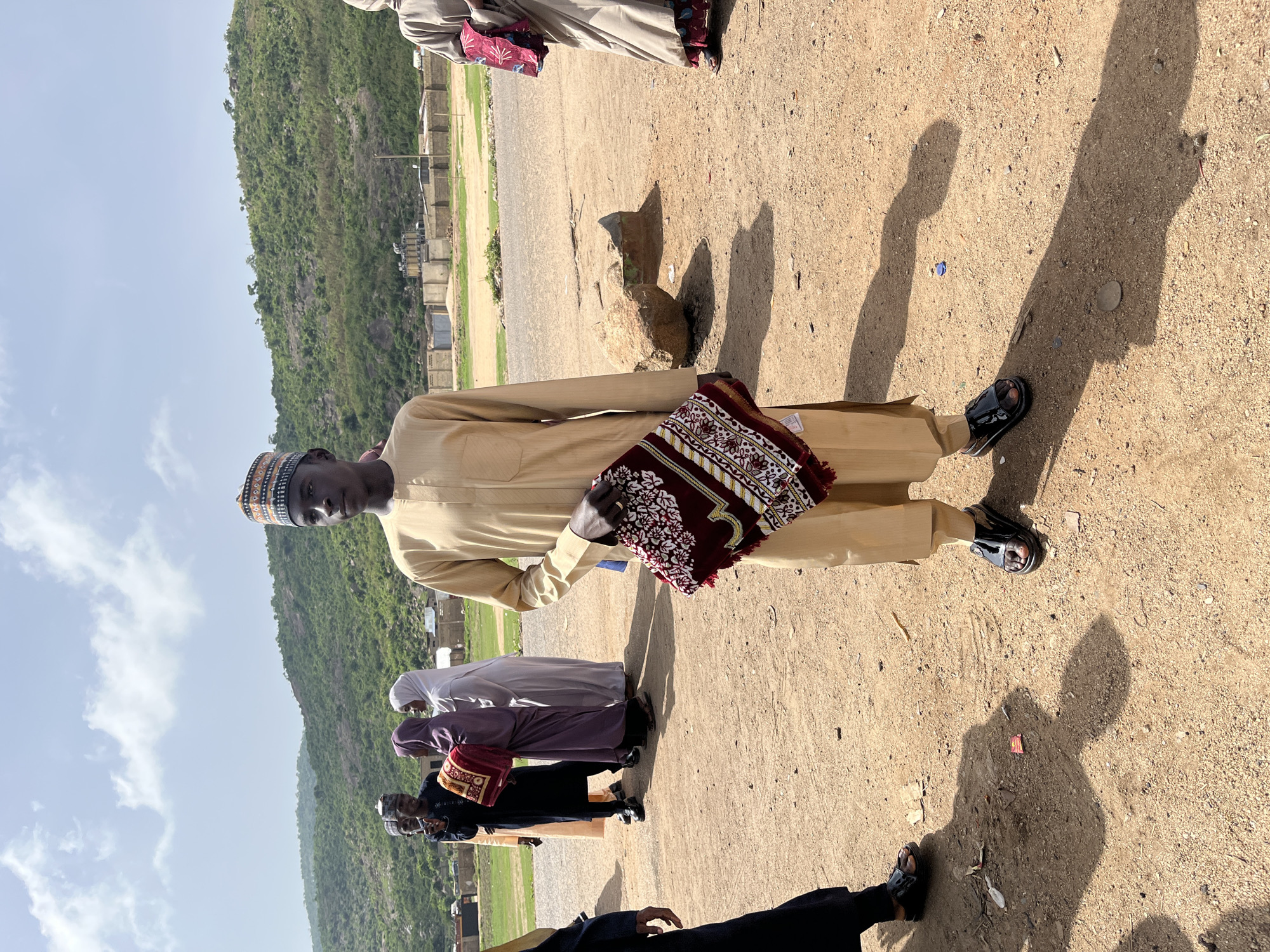 In Naraguta Hausawa, Nigeria, a group of people are outdoors on a dirt ground with sparse vegetation. A man stands in the foreground wearing a beige outfit, including a cap and sandals, holding a patterned bag. Behind him, other individuals are seen dressed in clothing that includes a hijab, robes, and carrying similar patterned bags. In the background, there is a hillside. Fences and box-like structures indicate a built environment on the flat land between the group and the hill. The sky is visible with some clouds. Shadows from people and objects are cast on the ground.