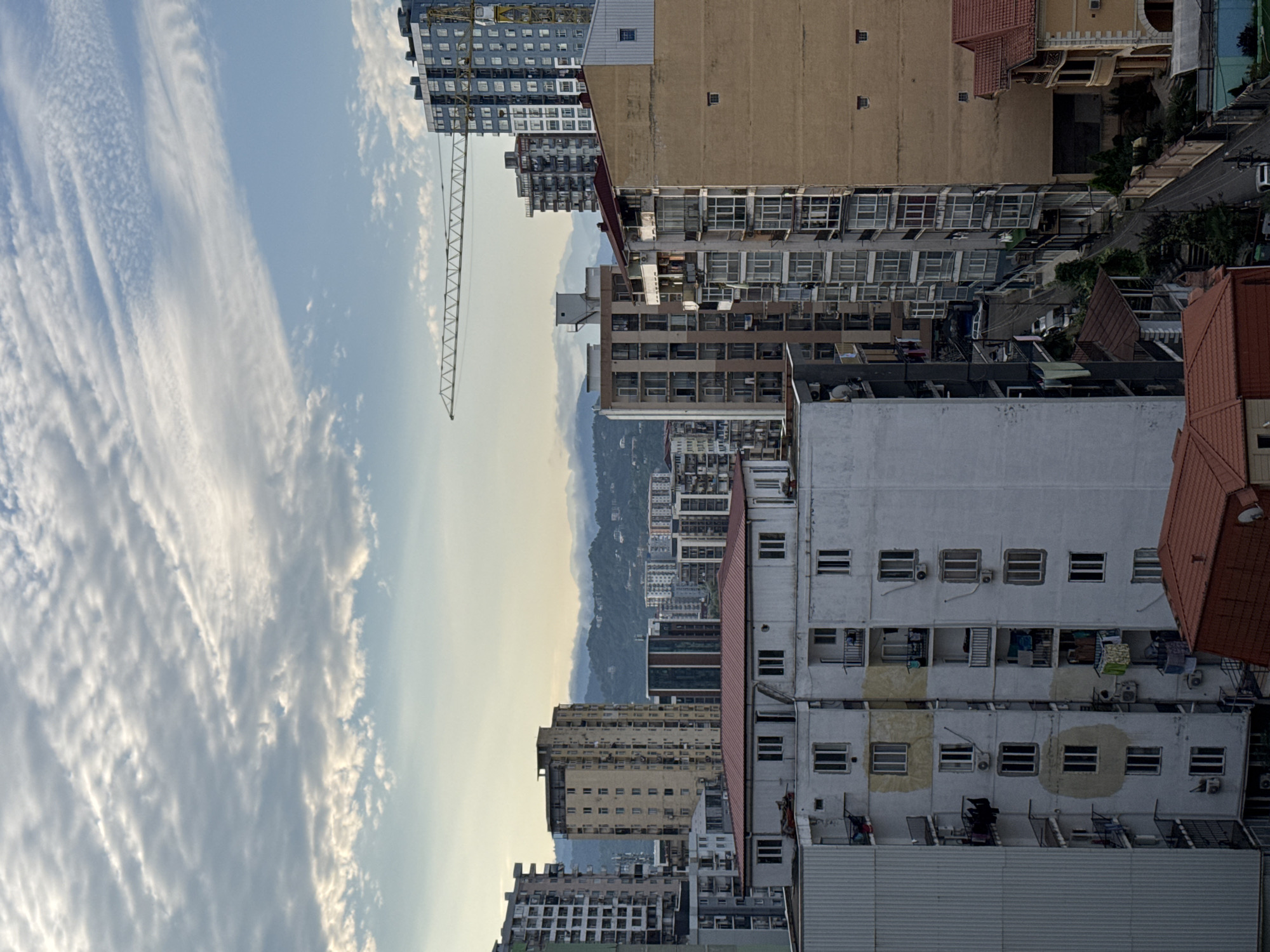 An elevated, eye-level, full outdoor shot captures a densely populated urban cityscape with a view of a mountainous horizon under a partially cloudy sky. In the foreground, several multi-story apartment buildings are prominently featured. The closest building on the left is a pale gray structure with vertical ribbed siding, partially obscuring a white apartment building with visible windows and balconies. Further back, more apartment buildings of varying heights and architectural styles dominate the scene, some appearing older and weathered, others modern and glass-fronted. A prominent crane, likely part of a construction project, stretches horizontally across the mid-ground, with a tall, blue-toned skyscraper visible to its right. The buildings are densely packed, suggesting a high-density urban environment. In the distance, rolling hills or mountains are shrouded in a light haze, blending into the pale sky. The overall impression is of a bustling city with ongoing development, common in areas like Batumi, Georgia, known for its coastal development and mountainous backdrop. No individuals are visible in the frame. The lighting suggests it is either early morning or late afternoon.