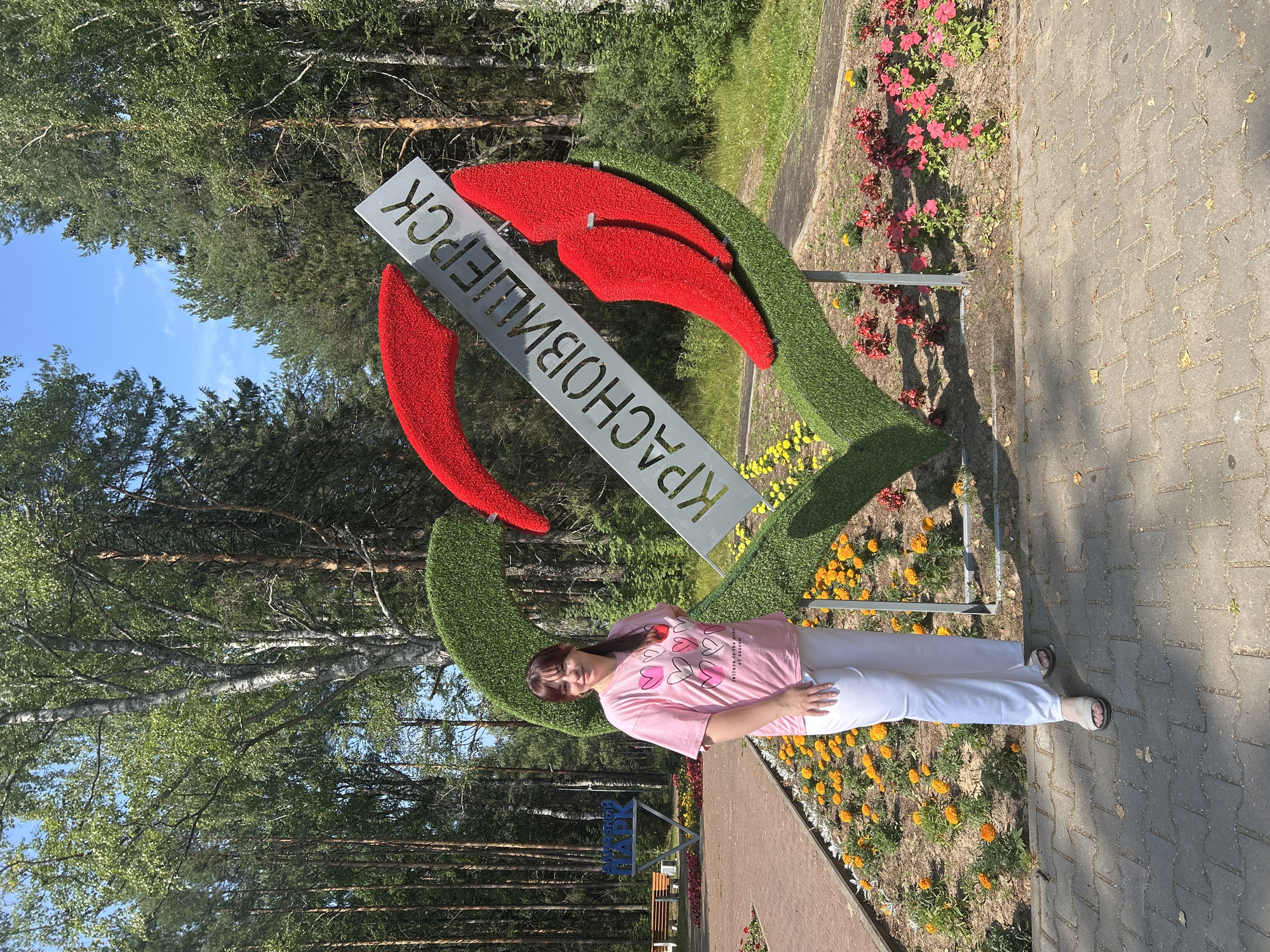In the city of Krasnovishersk, Russia, a woman stands in front of a large heart-shaped sculpture made of green artificial grass and red floral accents. A metal sign with Cyrillic lettering reading "КРАСНОВИШЕРСК" is attached to the sculpture. The woman is wearing a pink t-shirt with multiple heart designs and white pants. She is posing with her hand on her hip. The sculpture is situated in a park-like setting with trees in the background. A pathway paved with grey bricks is in the foreground, and flowerbeds with yellow and red flowers surround the sculpture. A sign that reads "МЕДНЫЙ ПАРК" is visible in the background to the left.
