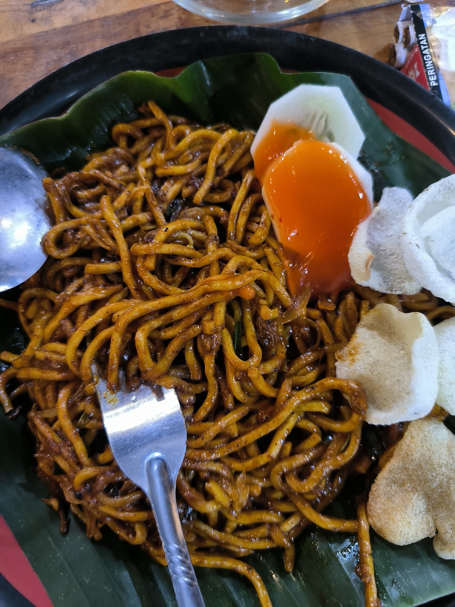 A close-up shot depicts a plate of Mie Aceh, an Acehnese spicy noodle dish, in Cempeudak, Indonesia. The dish features thick yellow noodles coated in a dark, rich sauce, garnished with slices of cucumber, a dollop of chili sauce, and crispy white crackers. A silver spoon and fork rest on the noodles, which are arranged on a banana leaf lining the plate. A glimpse of a wooden table and a condiment packet are visible in the background.