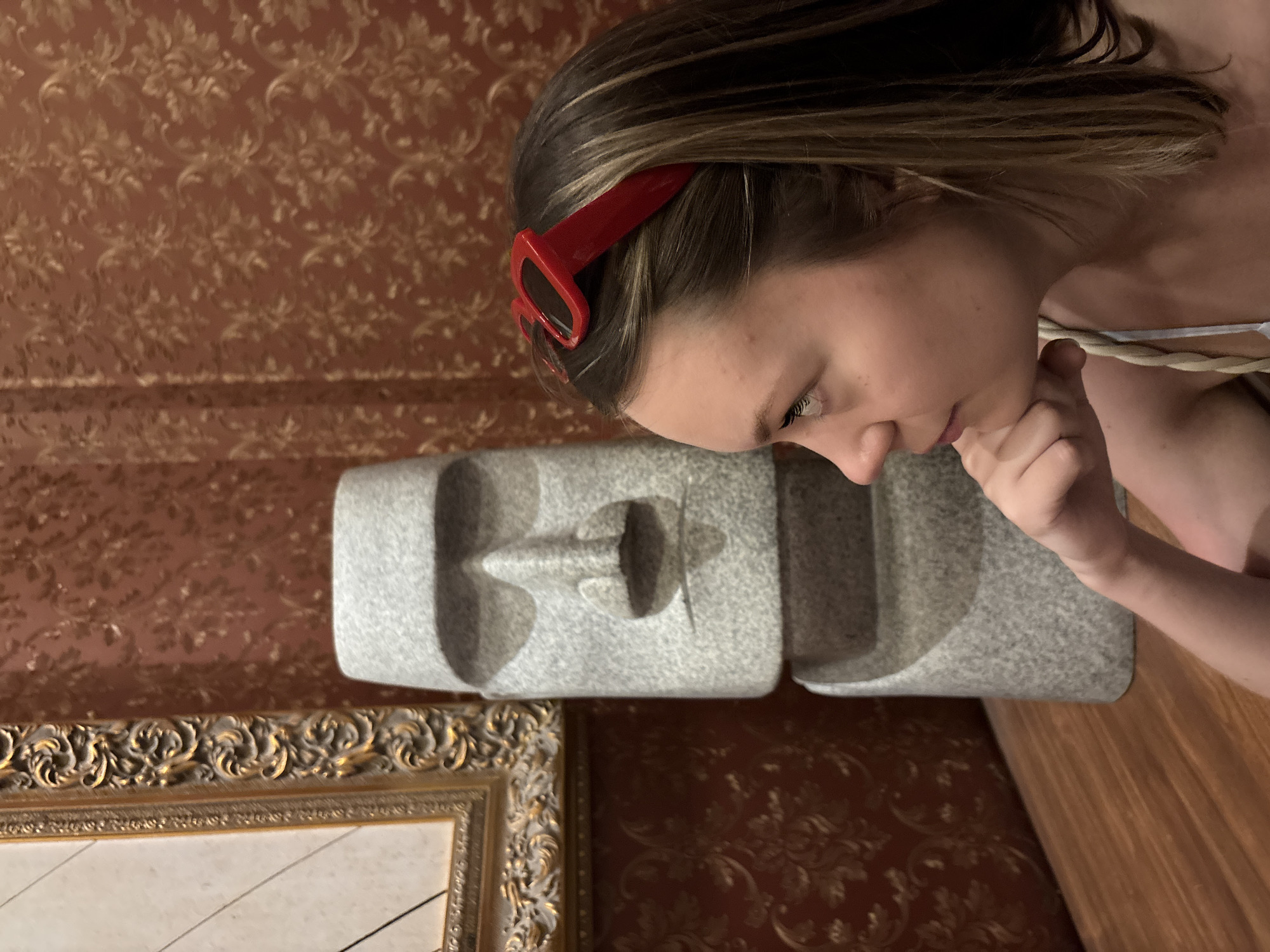 An indoor scene captures a young person intently observing a grey stone sculpture against a richly patterned wall. The person, with light brown hair and wearing red sunglasses atop their head, looks down at the object with their right hand near their chin, suggesting contemplation or curiosity. They are wearing a light-colored, thin-strapped top.
The grey sculpture, made of a granular, possibly granite-like material, appears as two sections. It features a stylized, abstract face with a prominent nose and hollowed-out features, resembling a mask or an ancient figure.
The background wall is covered in a reddish-brown wallpaper adorned with an intricate, repeating floral or damask pattern in a lighter, golden-brown hue. To the lower left, a portion of an ornate golden picture frame with elaborate, baroque-style carvings is visible. A wooden surface, possibly a floor or furniture, is present on the right side of the image. The setting suggests an environment with artistic or traditional decor, potentially located in Nha Trang Ward, Vietnam. No text is visible.