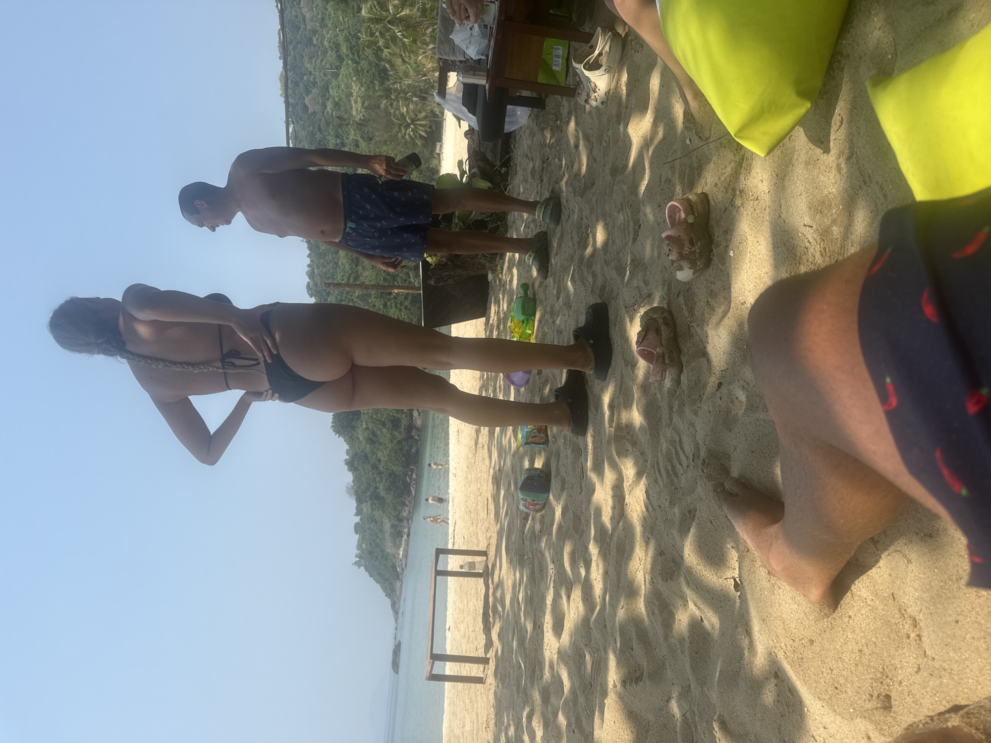 The image shows a beach scene. In the foreground, there is a leg in swim trunks with a chili pepper pattern. Next to it are two yellow beach cushions, and some pink kids' sandals. Behind these items, a partially obscured table is visible. Further back, a woman wearing a black bikini and black shoes stands with her back to the camera. To her right stands a man with blue swim trunks. The beach extends towards the sea, where people are visible in the water. A metal structure is set up in the sand closer to the water. The background shows a lush, green coastline. The sky is blue and clear. Đà Nẵng, Vietnam, is not specifically identifiable in the scene.