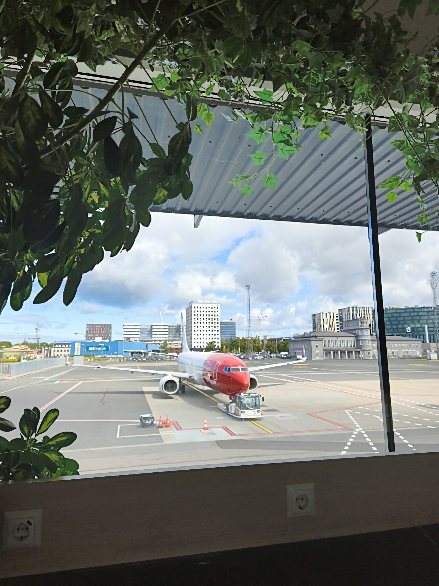 The image captures a view of Tallinn Airport (TLL) in Tallinn, Estonia, seen through a window adorned with artificial foliage. In the foreground, an electrical outlet is visible on a textured wall. The main subject of the image is a red and white airplane, possibly a Boeing 737, parked on the tarmac. A pushback tractor is connected to the aircraft's nose. The tarmac is marked with yellow and white lines, and several orange traffic cones are strategically placed. Beyond the tarmac, a row of modern buildings with glass facades and distinctive architectural styles are visible, including one with the text "Ulemiste City" on it. The sky is partly cloudy, with a mix of blue sky and white clouds. The overall scene depicts a typical airport environment on a bright day.