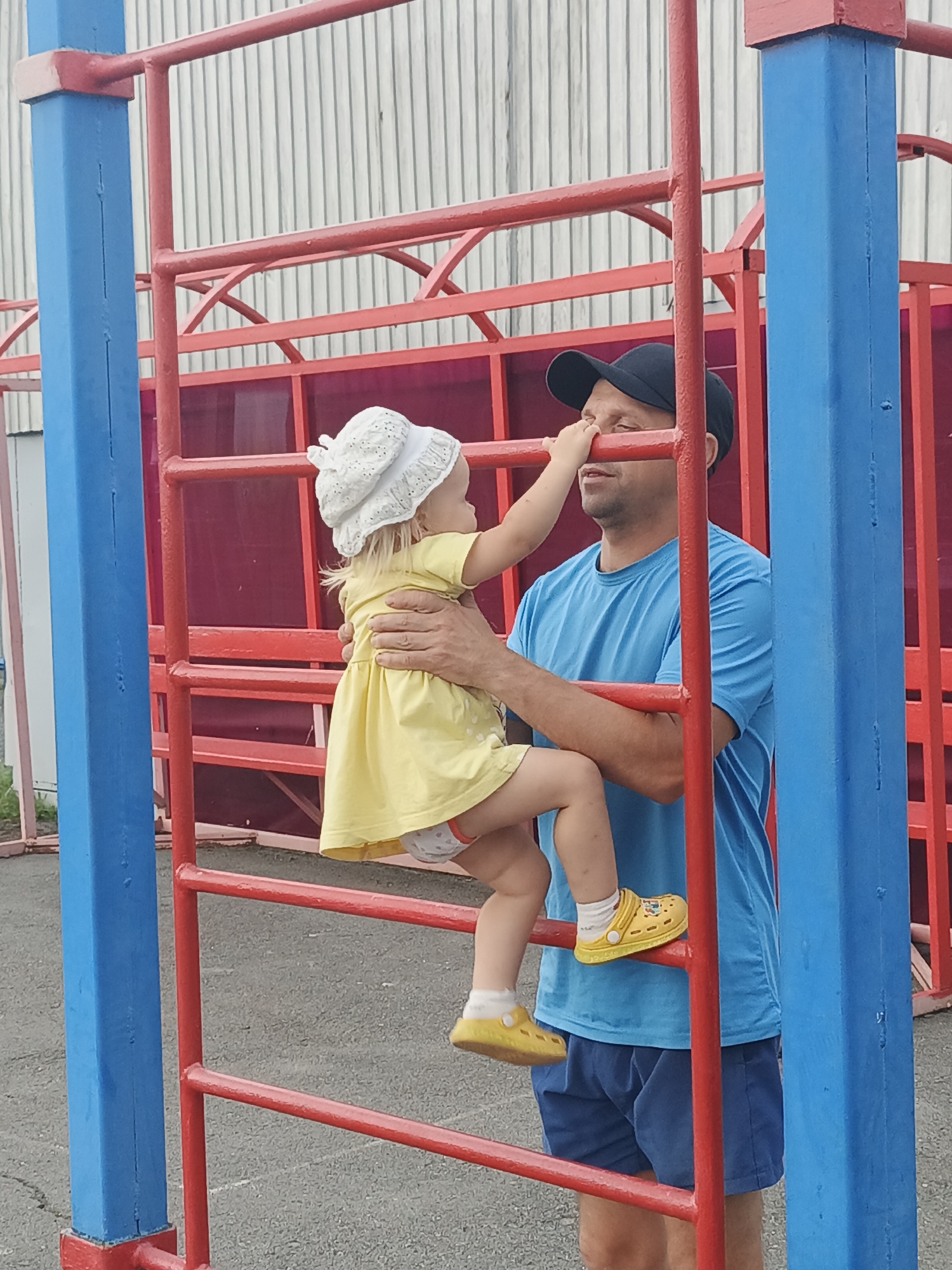 In Zlatoust, Russia, a man in a blue t-shirt and blue shorts, wearing a black baseball cap, is standing next to a red and blue jungle gym. He is holding a young child who is wearing a yellow dress and a white hat. The child is reaching up towards the man's face. The jungle gym is made of metal and has red bars. The background is a white building with vertical siding. The ground is asphalt.