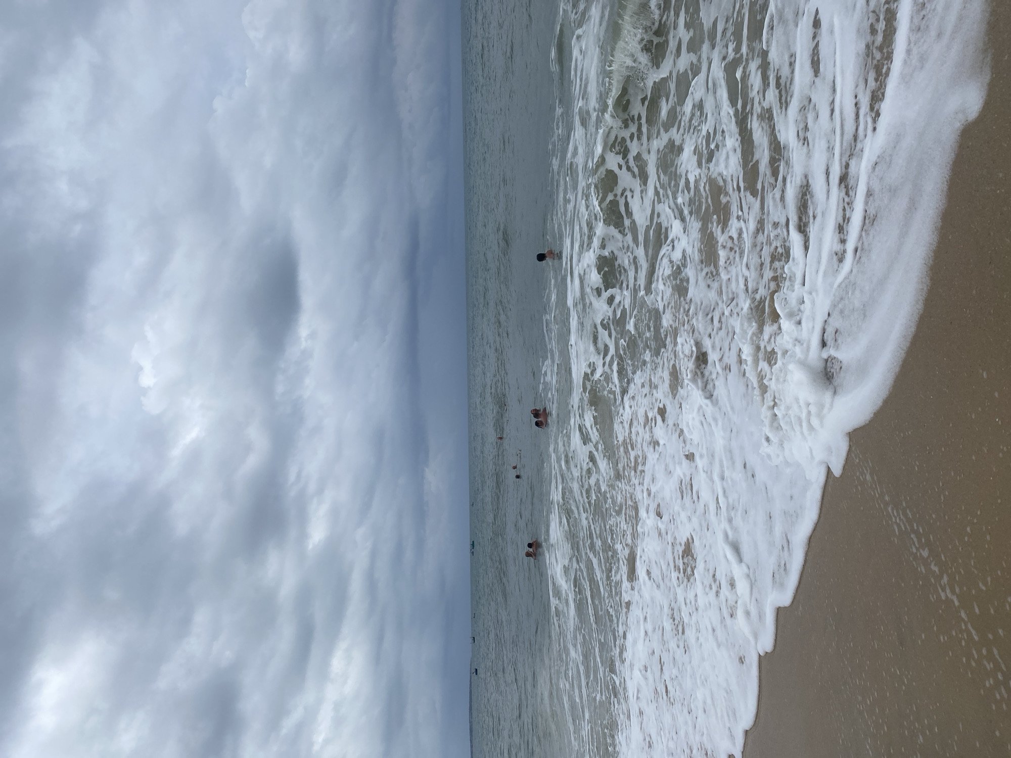 The image displays a coastal scene on an overcast day, captured from a low angle at the water's edge. The sky, occupying the upper portion of the frame (when correctly oriented), is filled with dense, grey clouds, indicating cloudy weather and diffused light.
The majority of the image is dominated by the ocean, which appears a muted grey-green under the cloudy sky. Several individuals are visible in the water, appearing to be swimming or wading. They are scattered across the mid-ground, some closer to the shore and others further out. Their forms are largely indistinct due to distance. A small, dark object, possibly a boat or buoy, is faintly visible on the distant horizon line.
In the foreground, foamy white waves are breaking and receding onto a light brown sandy beach. The foam creates dynamic patterns as it spreads across the wet sand. The scene suggests a calm, leisure activity, likely during the daytime, despite the lack of direct sunlight. No visible text is present. The setting is a beach, consistent with the provided location context of Phu Thuy Ward, Vietnam.