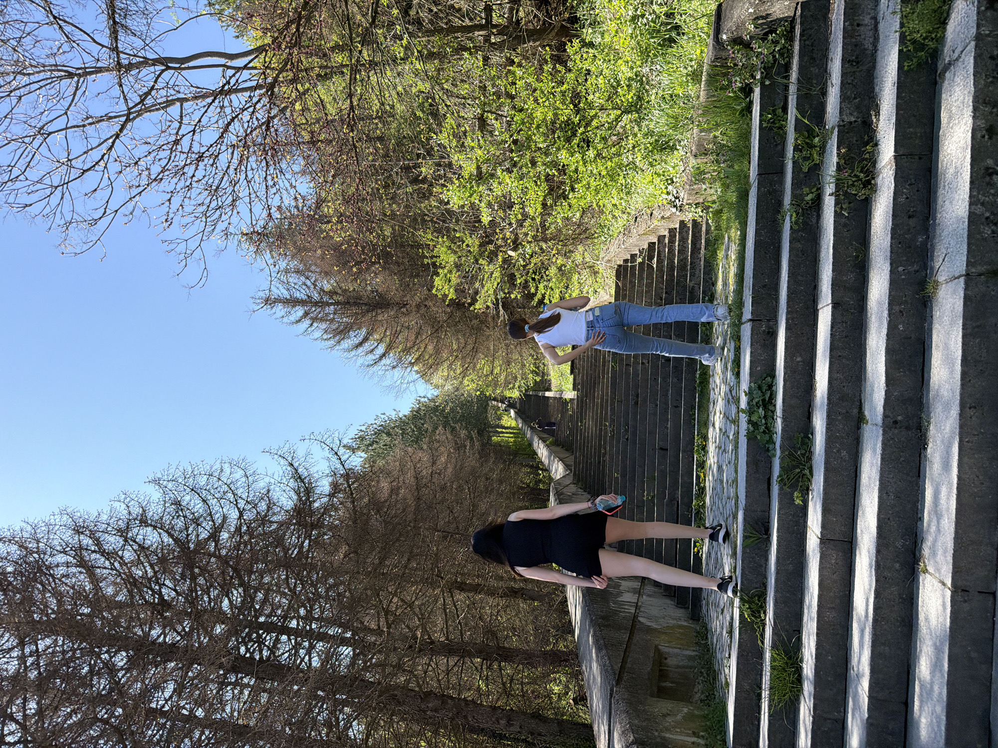 Two women are on a long set of stone stairs that extend into the distance, lined on either side with trees. One woman, closer to the camera, is wearing a black dress and black and white shoes, and is carrying a blue water bottle in her left hand. The other woman, further up the stairs, is wearing a white shirt and blue jeans. The scene is set in a park in the city of Shumen, Bulgaria. The sky is blue, and the trees are a mix of bare branches and green leaves.