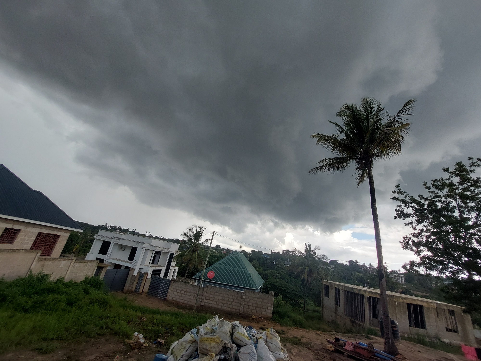 The image presents a landscape dominated by a heavily overcast, dark grey sky, indicating an impending storm or recent rainfall. Voluminous, dense clouds stretch across the upper two-thirds of the frame, casting a dim light over the scene.
Several buildings and structures are visible across the frame. On the far left, part of a house with a dark roof and light-colored, possibly unfinished, walls featuring reddish-brown window grates can be seen. Further back on the left, a multi-story white building with dark windows and an external air conditioning unit is partially obscured by a fence. In the mid-ground, a house with a distinctive bright green pyramidal roof and a red satellite dish is situated behind a concrete block wall. On the right, a tall, slender palm tree stands prominently, with an unfinished concrete structure, possibly a building under construction, located behind it on a sloping terrain.
The foreground features a pile of white and tan sacks, some with partially visible text such as "cement" or "storage cement," alongside blue containers and other debris on bare earth and sparse green vegetation. Lush green foliage, including other palm trees, covers the hills in the background, where additional residential buildings are scattered.
No people are visible in the image. The scene depicts a quiet, developing residential area experiencing significant cloud cover, suggesting a moody atmosphere. The architecture, tropical vegetation, and cloudy weather are consistent with a location like Dar es-Salaam, Tanzania.