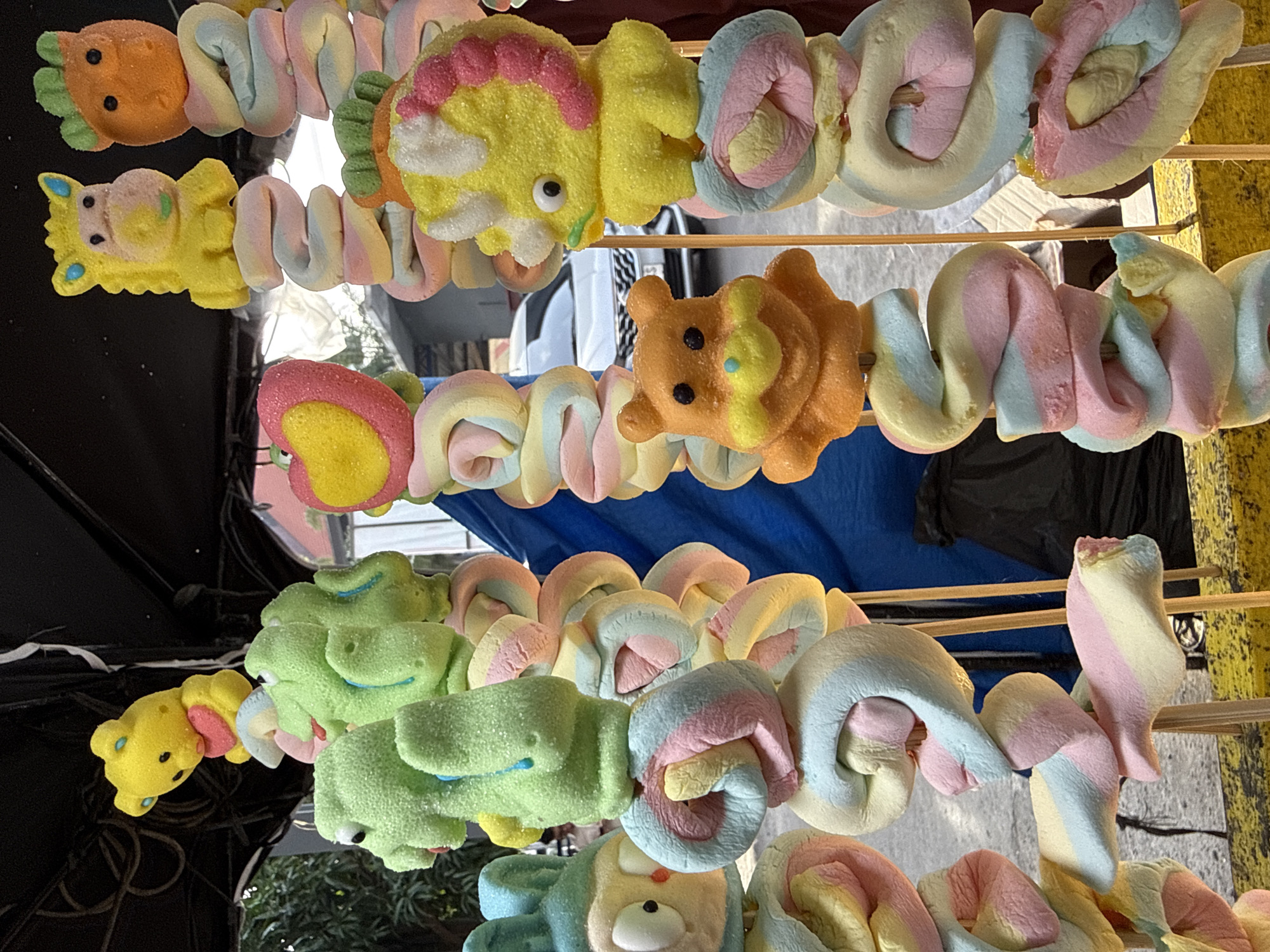 In Lhokseumawe, Indonesia, a close-up shot captures an array of colorful, character-shaped marshmallows skewered on sticks. The marshmallows, which include teddy bears, frogs, a bunny, a horse, a dinosaur, and other animal figures, are coated in a light dusting of sugar and arranged vertically. Interspersed between the character marshmallows are twisted, pastel-colored marshmallow segments in shades of pink, yellow, and blue, also threaded onto wooden skewers. The background reveals a blurry street scene, with a white car visible in the middle ground and a dark blue tarp behind the marshmallow display. The foreground is dominated by a yellow surface, possibly a table or cart, on which the skewers rest. The overall impression is a vibrant and whimsical street food vendor display.