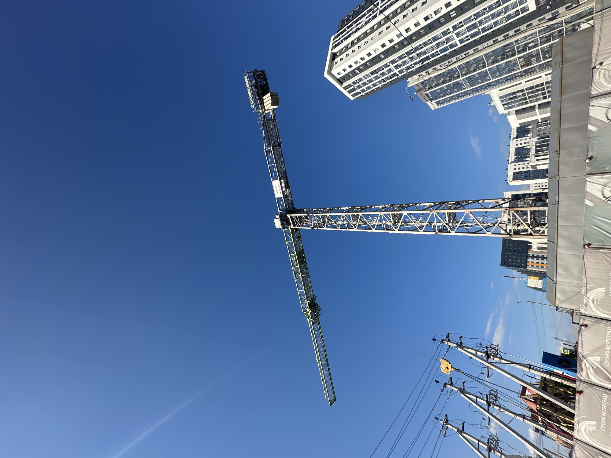 A low-angle, wide shot captures a construction scene under a clear blue sky with a single white construction crane dominating the frame. The crane, with its long horizontal jib, extends from the right side of the image into the left. Beneath the crane and to the right, a modern, white and grey apartment building with numerous windows is visible, tilted at an angle due to the camera's perspective. Several other multi-story buildings, some appearing lighter in color with similar window patterns, are clustered behind the main apartment building. In the foreground, to the left, a series of utility poles with electrical wires are positioned at an angle. The base of the construction crane is partially obscured by a grey construction barrier with a repeating circular pattern and text that is partially visible. The overall scene depicts an active urban construction site.