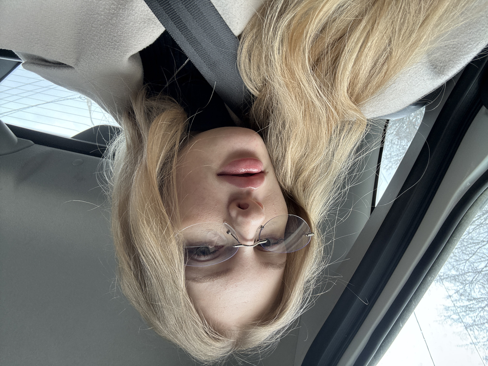 A young woman with blonde hair and rimless glasses is sitting in a car, wearing a beige coat and a black top, and has a seatbelt on. The photo appears to have been taken in Perm, Russia, as determined based on the visible objects in the window and the general background environment.