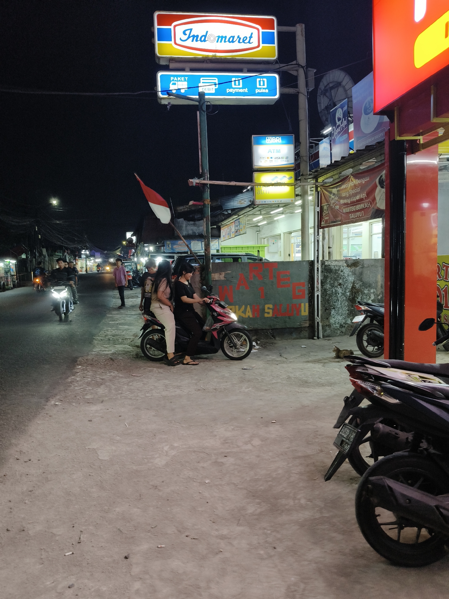 The image depicts a street scene at night in South Tangerang, Indonesia, likely on Jalan Raya Pahlawan. An Indomaret convenience store sign is prominently displayed, featuring its logo and smaller signs indicating services like "PAKET," "DEBIT," and "PULSA." Adjacent to this, a red and yellow illuminated sign can be seen. A group of people are present, with two individuals on a pink and black scooter in the foreground. Another scooter is parked nearby. In the background, several other motorcycles and individuals are visible on the street. The environment is a typical urban street at night, with streetlights illuminating the scene and a building that appears to be a "WARTEG" (a type of Indonesian food stall) with a sign reading "WARTEG BERKAH SALUYU 1" and "AKAN SALUYU". Indonesian flags are also visible, adding a cultural element to the scene. The overall impression is that of a busy commercial street at night.