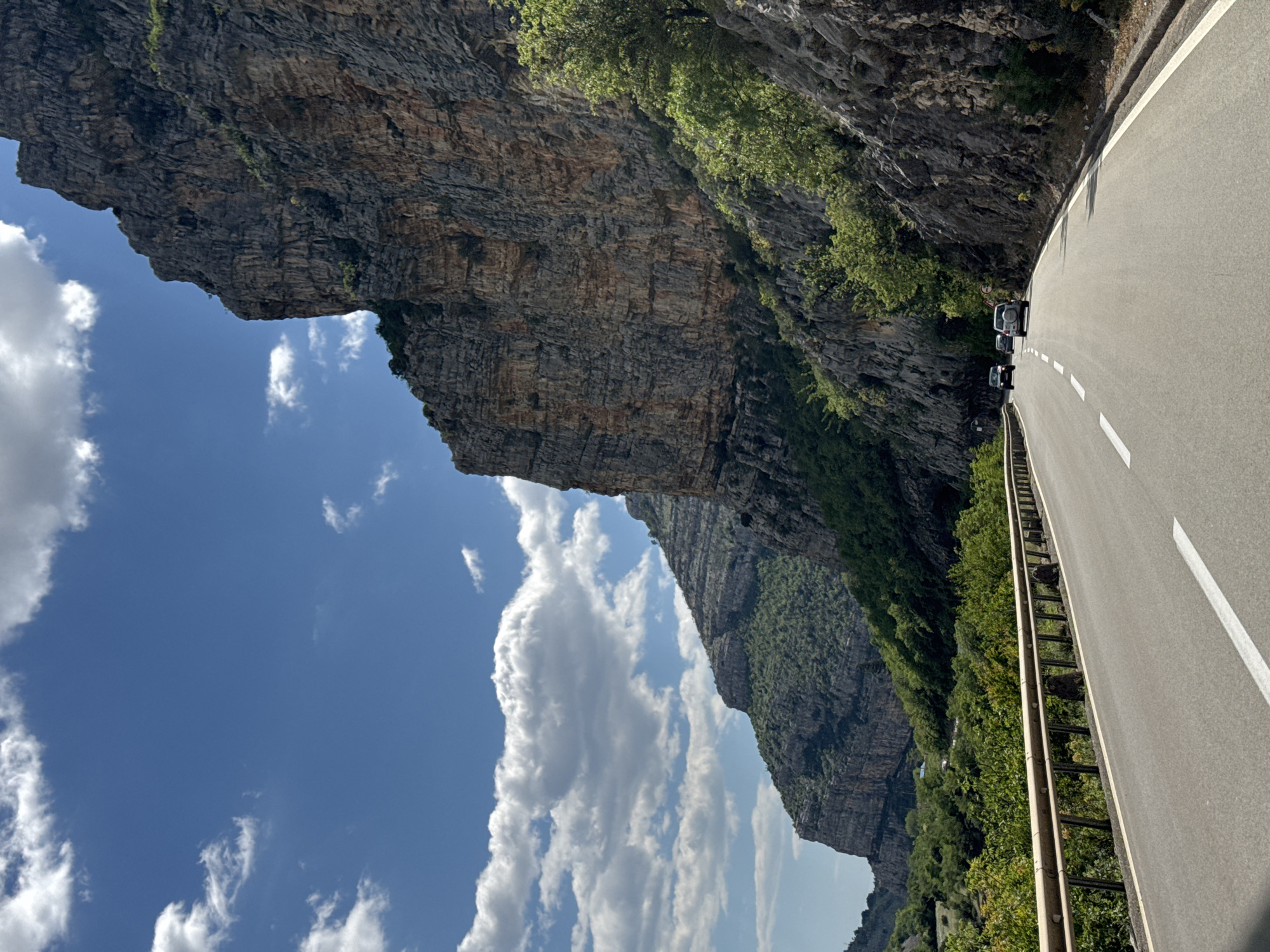 The image depicts a road in Donje Mrke, Montenegro, winding through a narrow gorge. A sheer rock face rises on the left side of the road, contrasting with a blue sky dotted with white clouds. Vegetation clings to the rock face in places. On the right side of the road, a metal guardrail separates the road from a steep drop-off covered in dense greenery. Several cars are parked on the side of the road.