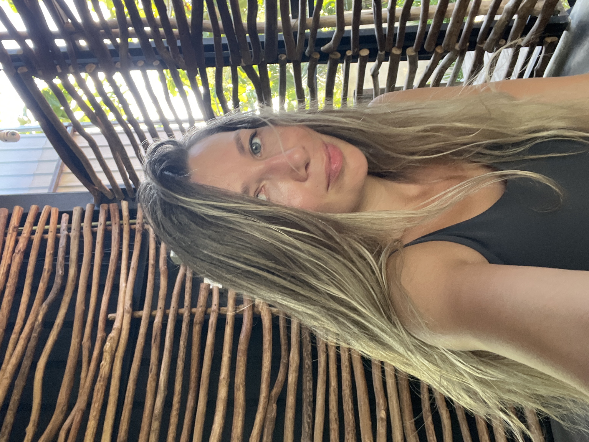 A woman with long, blonde, wavy hair is shown in a close-up, shoulder-up shot. She is wearing a dark grey tank top and looking to her left with a slight smile. Her left eye is partially closed. The background consists of a wooden structure made of horizontally laid branches, which creates a textured pattern. Sunlight streams through the gaps in the wood, illuminating the scene and casting shadows. The environment appears to be outdoors, possibly a resort or cafe in Weligama, Sri Lanka, suggested by the natural wood elements and tropical vegetation visible through the structure.