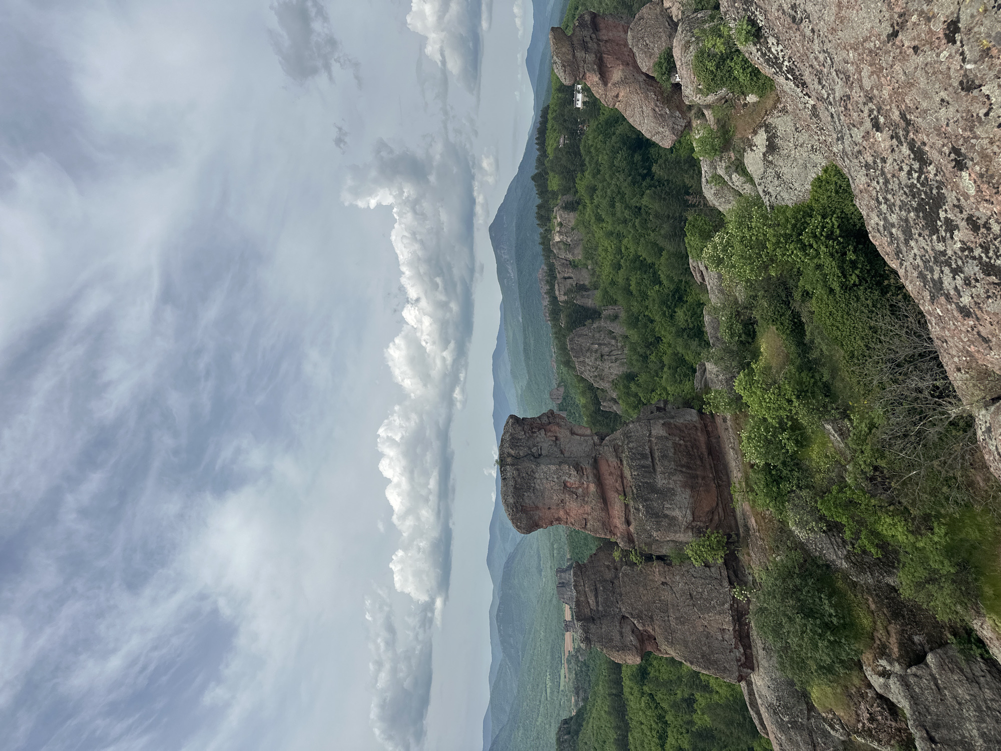 The image shows a landscape in Belogradchik, Bulgaria, featuring sandstone rock formations covered in green vegetation. The sky is filled with clouds. There are forested mountains in the background. The landscape appears to be a natural attraction or park.