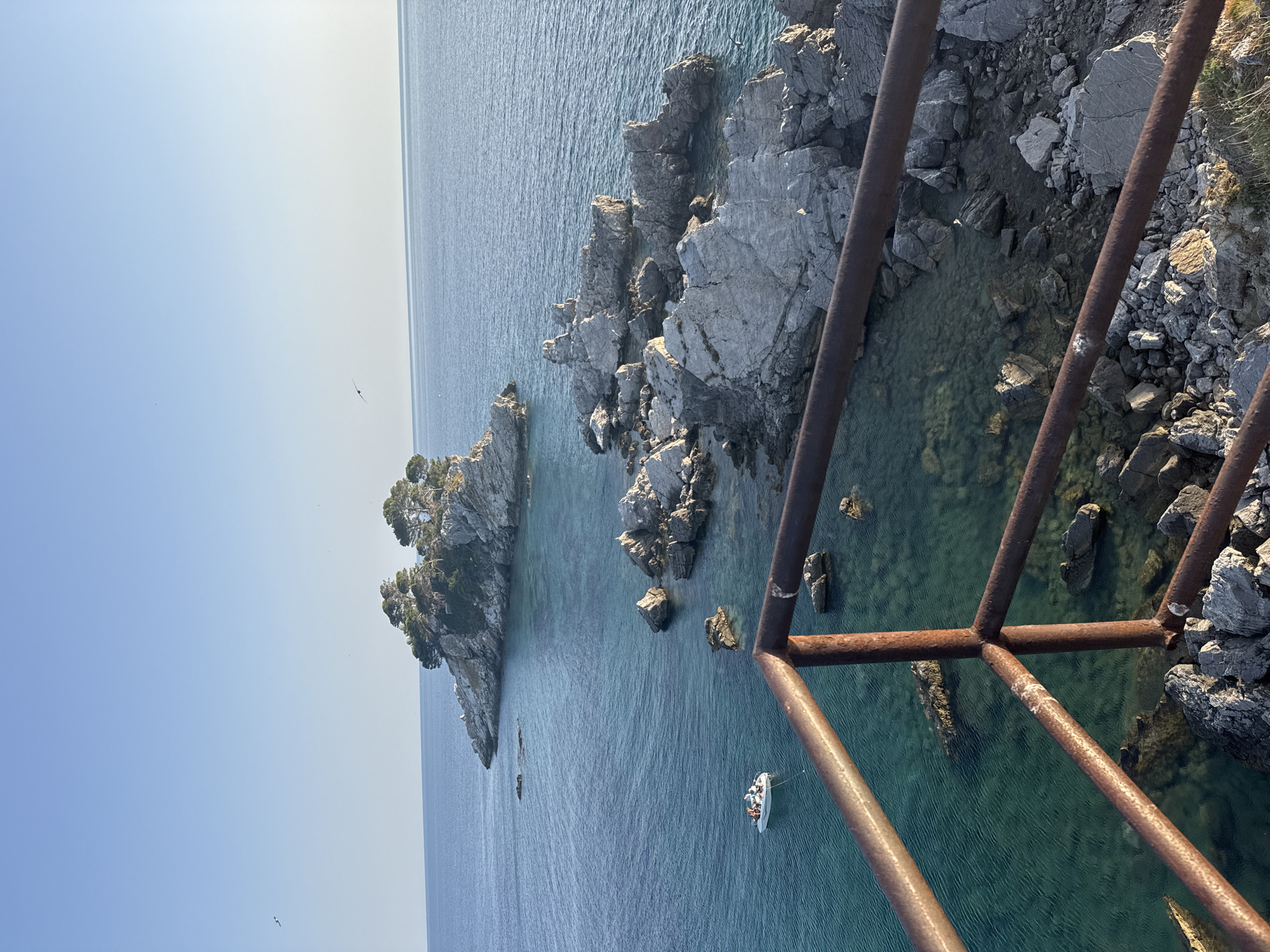 A scenic view of the Adriatic Sea coastline near Petrovac centar, Montenegro, captures a small, rocky island topped with trees in the distance. In the foreground, a rusty metal railing partially obstructs the view of the turquoise water, which reveals submerged rocks. A small white boat with several people onboard is visible on the water. The clear blue sky and the calm sea suggest a peaceful day.