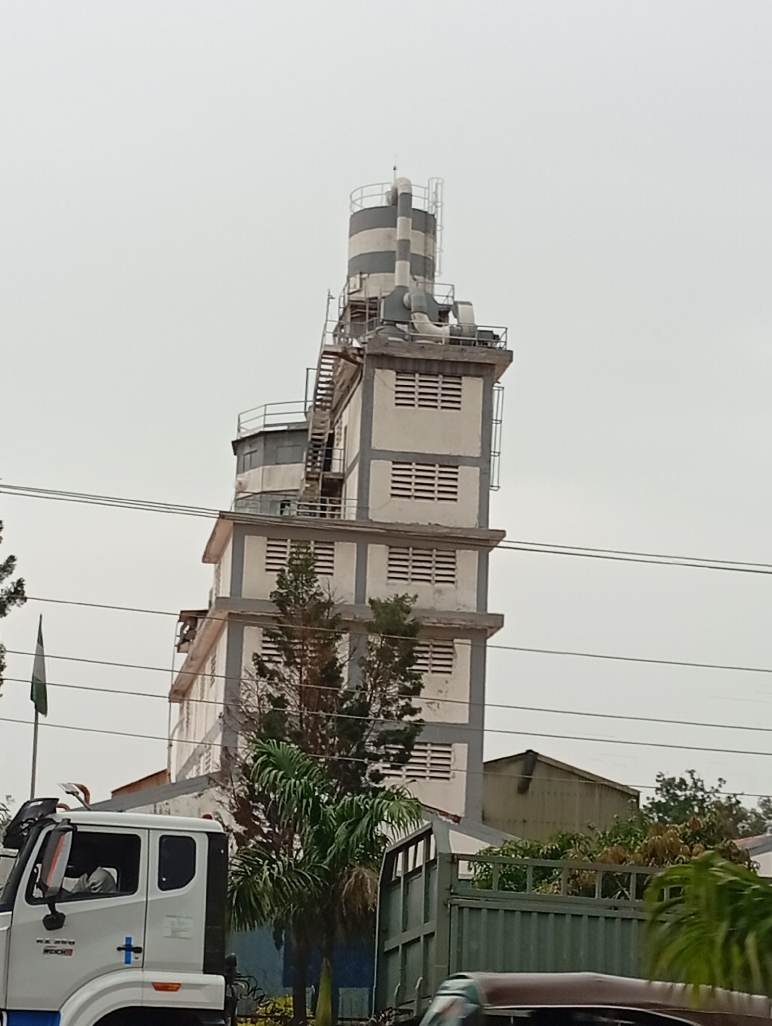 An industrial tower, painted in alternating gray and white horizontal stripes, dominates the center of the frame. The tower features multiple levels with rectangular, louvered windows and external staircases and pipework. To the left of the tower, a white truck with a blue cross logo is partially visible. A flag, green and white, flies on a pole near the truck. Lush green trees and foliage are scattered around the base of the tower and in the foreground. The scene is set against a pale, overcast sky. The architectural style and the presence of the flag suggest a location in Nigeria, possibly Jos, given the context provided. The overall impression is of an industrial facility in an urban or semi-urban environment.