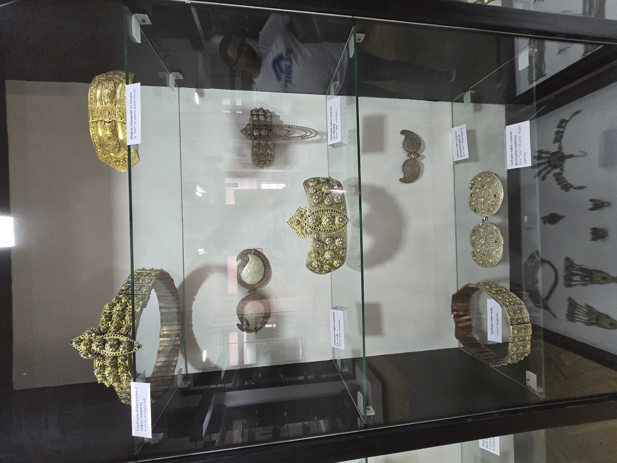 The image displays a large, vertical glass display case containing various historical metal artifacts, identified as belts and belt buckles ("pafti" or "chaprazi"), likely exhibited at a museum in Teteven, Bulgaria.
The display case features multiple glass shelves supported by dark vertical frames. The items are illuminated by overhead lighting, causing reflections on the glass.
On the top shelf, a large, ornate golden-yellow belt with an elaborate buckle ("A silver wrought belt") is displayed on the left, its segmented lower portion resting on the shelf. To the right, another golden-yellow, highly decorative belt buckle ("A "belt" of yellow metal (brass)") is positioned horizontally.
The middle shelf presents three smaller items. On the far left, a pair of oval silver buckles with white pearl-like centers ("Silver "belts" with pearl") are shown. In the center, a highly ornate, gilded buckle or belt segment adorned with numerous raised spherical elements and intricate filigree is placed. To its right, a silver buckle featuring multiple chains hanging from it ("Silver "belts" with chain") is mounted against the back of the case.
The third shelf from the top holds a silver wrought belt ("A silver wrought belt") on the left, similar in construction to the top left item but with smaller, rectangular decorated segments. To its right, a pair of small, wing-shaped filigree silver buckles ("Silver "belts" with filigree engraving") are displayed. Below these, two circular silver buckles with intricate filigree and what appear to be small embedded gems ("Silver "belts" with gems - filigree engraving") are positioned.
On the floor of the display case, several dark, intricate metal objects are visible. These include what resembles a complex metal hair ornament or headdress with multiple radiating elements. A partial label indicates "Пояса на Св. Георги" ("A gold pl...") suggesting another gold plaque or item related to St. George or St. Hristophor.
A male individual is visible in reflection on the right side of the glass display case. He is wearing a light-colored short-sleeved t-shirt with a blue graphic logo and dark trousers, and glasses. His reflection indicates he is standing to the right of the display case, observing its contents or in the immediate vicinity. The background behind the display case is a plain, light-colored wall.