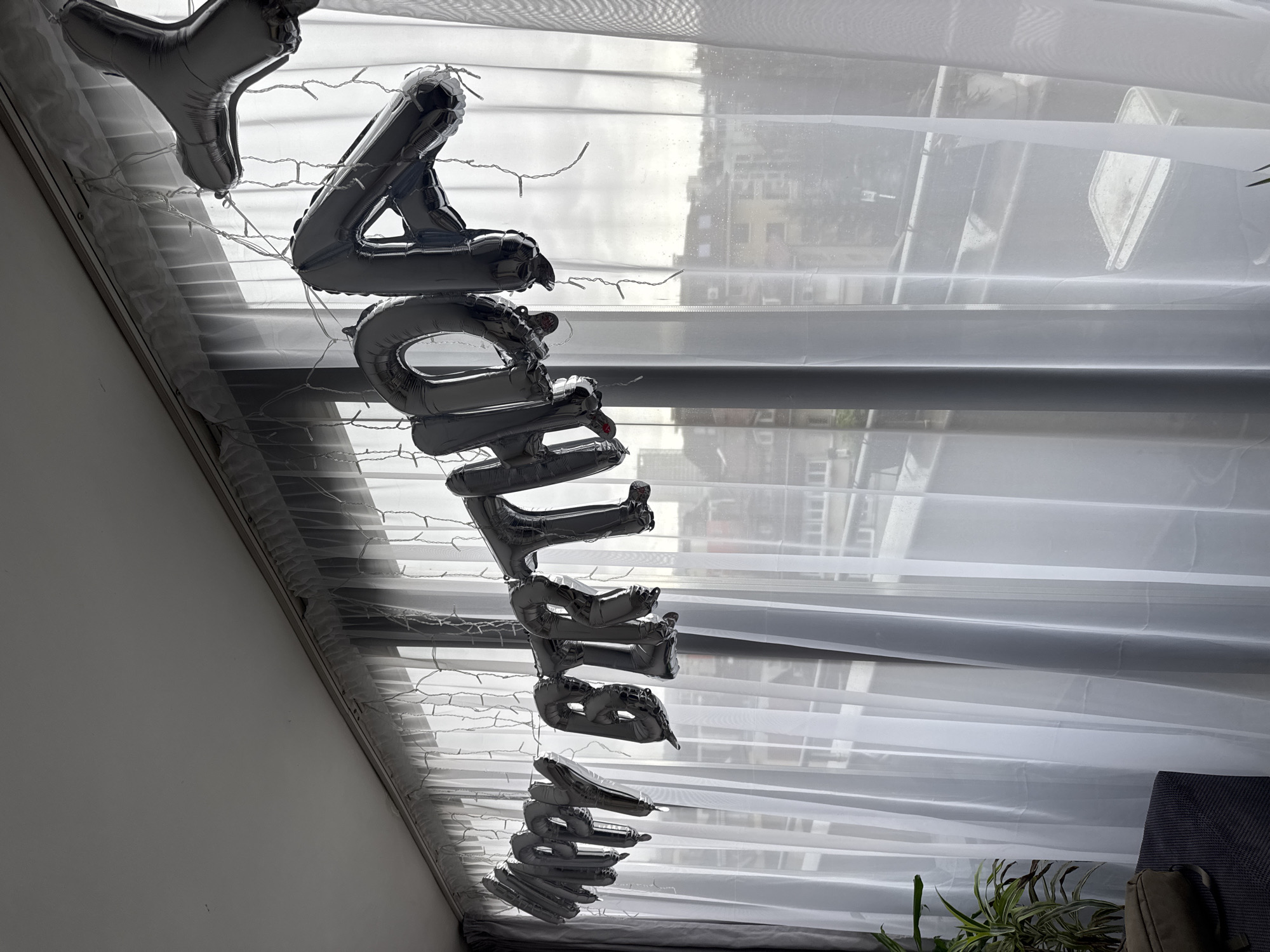 The image shows silver balloon letters spelling "birthday" hanging in front of a window. The view through the window shows a cityscape that is consistent with Sofia, Bulgaria. The balloons are attached to a string of fairy lights. A white sheer curtain hangs behind the balloons and in front of the window. Some greenery is visible in the lower right corner of the image.