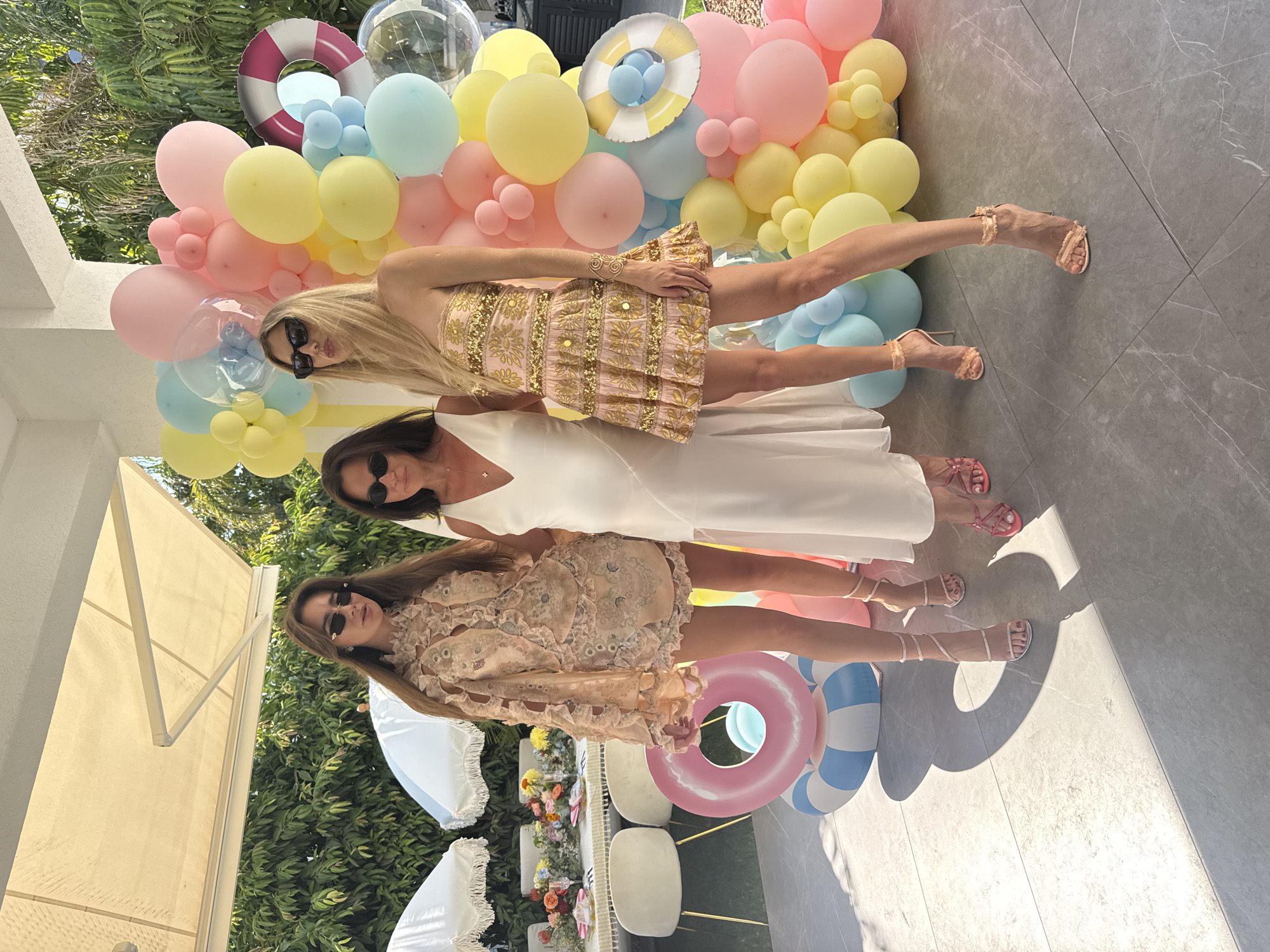 Three women are standing outdoors in front of a balloon arch and a table set for a party. The woman on the left is wearing a light pink, intricately detailed dress and sunglasses. The woman in the middle is wearing a white dress and sunglasses. The woman on the right is wearing a strapless pink and gold sequined dress and sunglasses. All three women are wearing high heels. In the background, there is a table laden with flowers and place settings, as well as umbrellas. A life preserver float and a large inflatable ring are also visible. The ground appears to be paved with large, grey tiles. The image suggests a party or celebration taking place outdoors, possibly by a pool or at a beachside venue, consistent with a location in Agios Tychona Community, Cyprus.