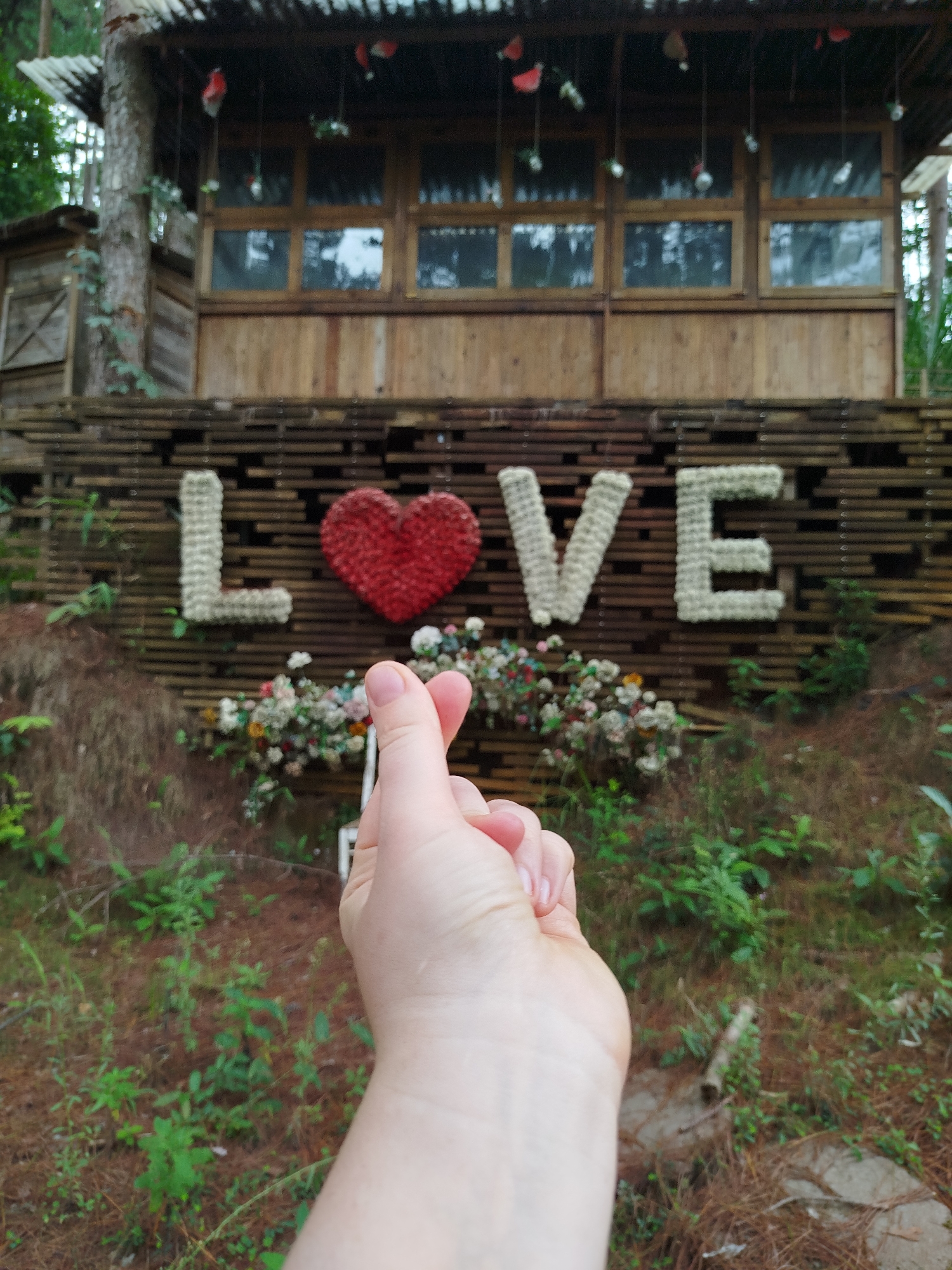 The image depicts a hand forming a heart shape in front of a decorative display that spells out "LOVE" with a red heart in place of the letter "O". The "LOVE" sign is made of white materials and is mounted on a wooden slatted background. Below the sign, there are floral arrangements. In the background, a rustic wooden building with multiple windows is visible, suggesting it could be a part of a resort or a themed attraction. The surrounding environment appears to be natural, with trees and vegetation, indicating an outdoor setting. The overall impression is of a romantic or recreational location, possibly in a rural or semi-rural area like Thôn 5, Vietnam, given the mention of the location.
