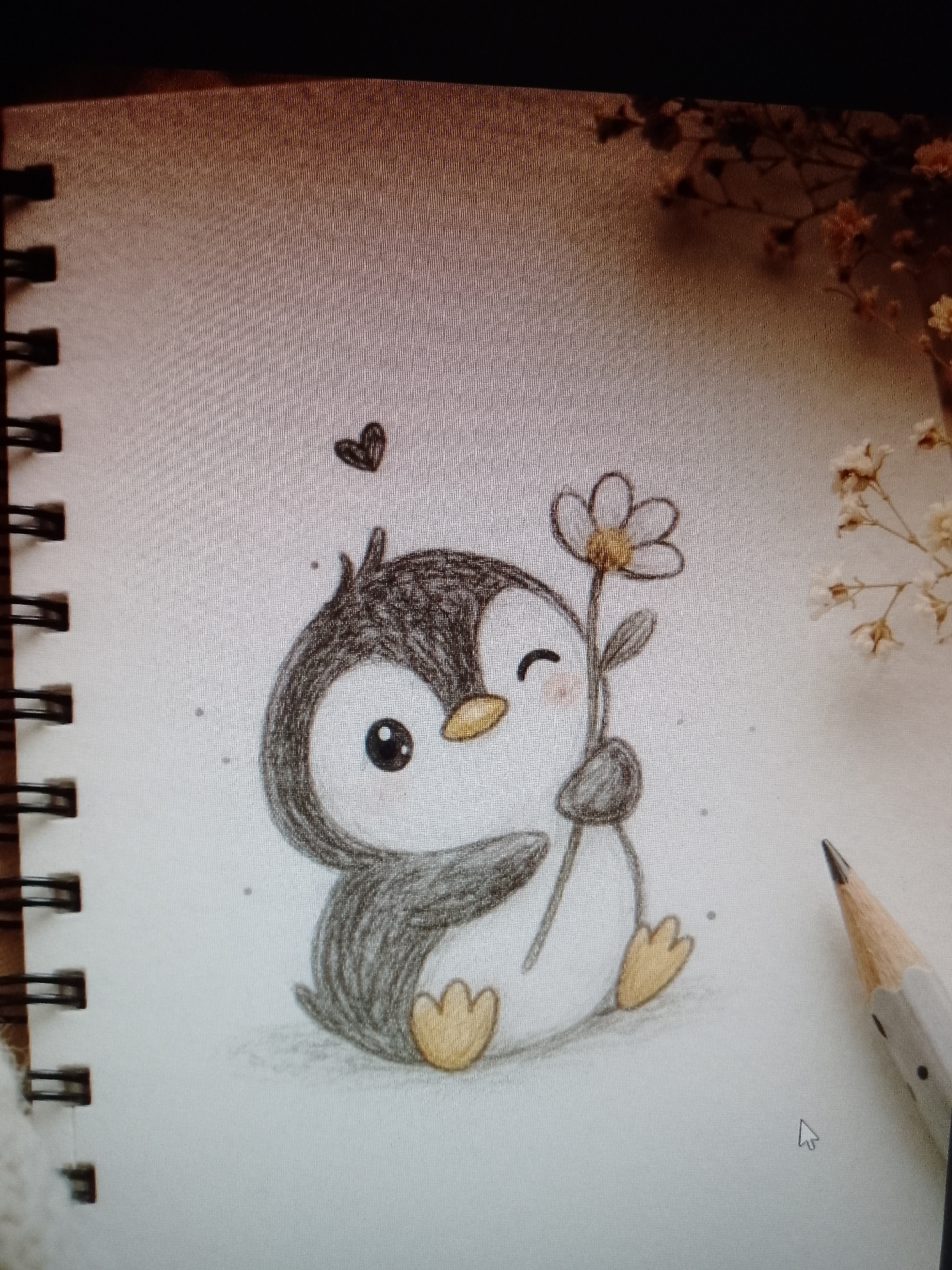 The image depicts a drawing of a cartoon penguin holding a daisy. The penguin is grey and white with yellow feet and a yellow beak, and a small heart is drawn above its head. The drawing is on a page in a spiral-bound notebook. A pencil is visible on the right side of the image, and dried flowers are in the background on the upper right.