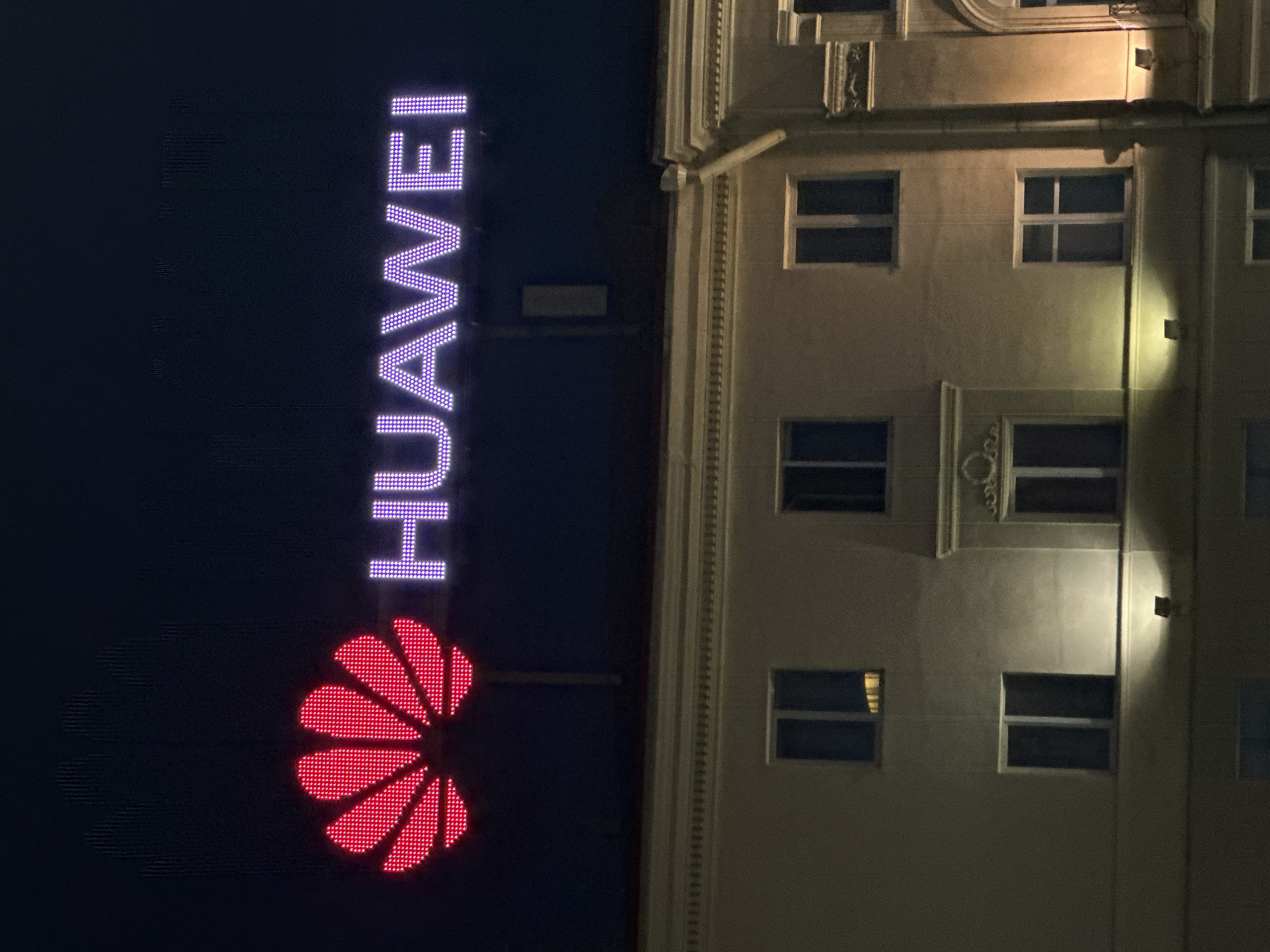An outdoor, medium shot captures a nighttime view of a building facade illuminated by a large, glowing "Huawei" logo positioned above it. The logo features a red flower-like symbol on the left and the word "HUAWEI" spelled out in purple LED lights to its right. The building below is a multi-story structure with several dark windows. The overall atmosphere is dark, with the illuminated logo being the most prominent feature against the night sky. The building's architecture appears to be classical, with decorative elements visible on the facade. The scene is devoid of people.