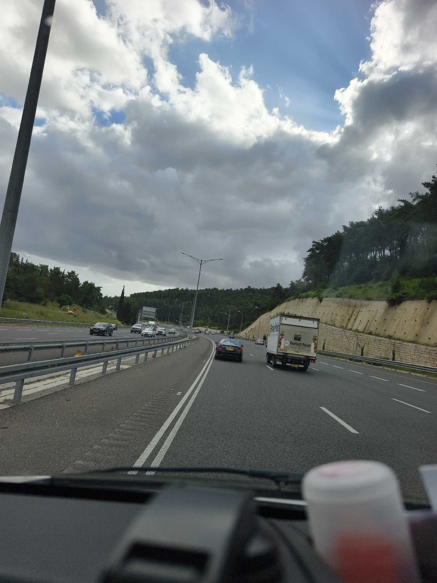 Captured from the interior of a moving vehicle on a highway near Yokneam Ilit, Israel, the image shows a multi-lane highway with other vehicles. The road is marked with solid and broken white lines. Guardrails line the side of the road. On the right, a hillside is covered with trees and a retaining wall. The sky is overcast with a mixture of gray and white clouds. In the distance, additional vehicles can be seen. A "Perfect Point" delivery truck is in the right lane. Parts of the vehicle's dashboard and a white, cylindrical object are visible in the foreground.