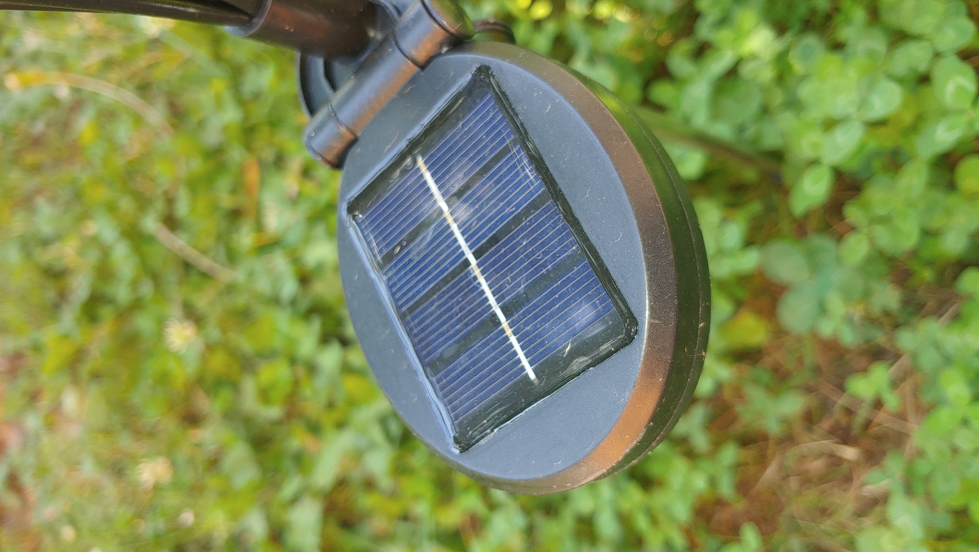A close-up, eye-level shot focuses on a small, oval-shaped solar panel on an outdoor light fixture. The solar panel, which is dark blue with thin, parallel lines, is set into a dark gray, circular casing. The casing is slightly raised at the edges and has a matte finish. To the right of the solar panel, a black metal support arm is visible, connecting the light fixture to an unseen pole or stake. The background is blurred and consists of green foliage, likely grass and low-lying plants, with some brown organic matter visible on the ground. The lighting suggests it is daytime, with sunlight reflecting off the solar panel. The scene appears to be outdoors in a natural or garden setting. No people or animals are present. The location is identified as Mukachevo, Ukraine.