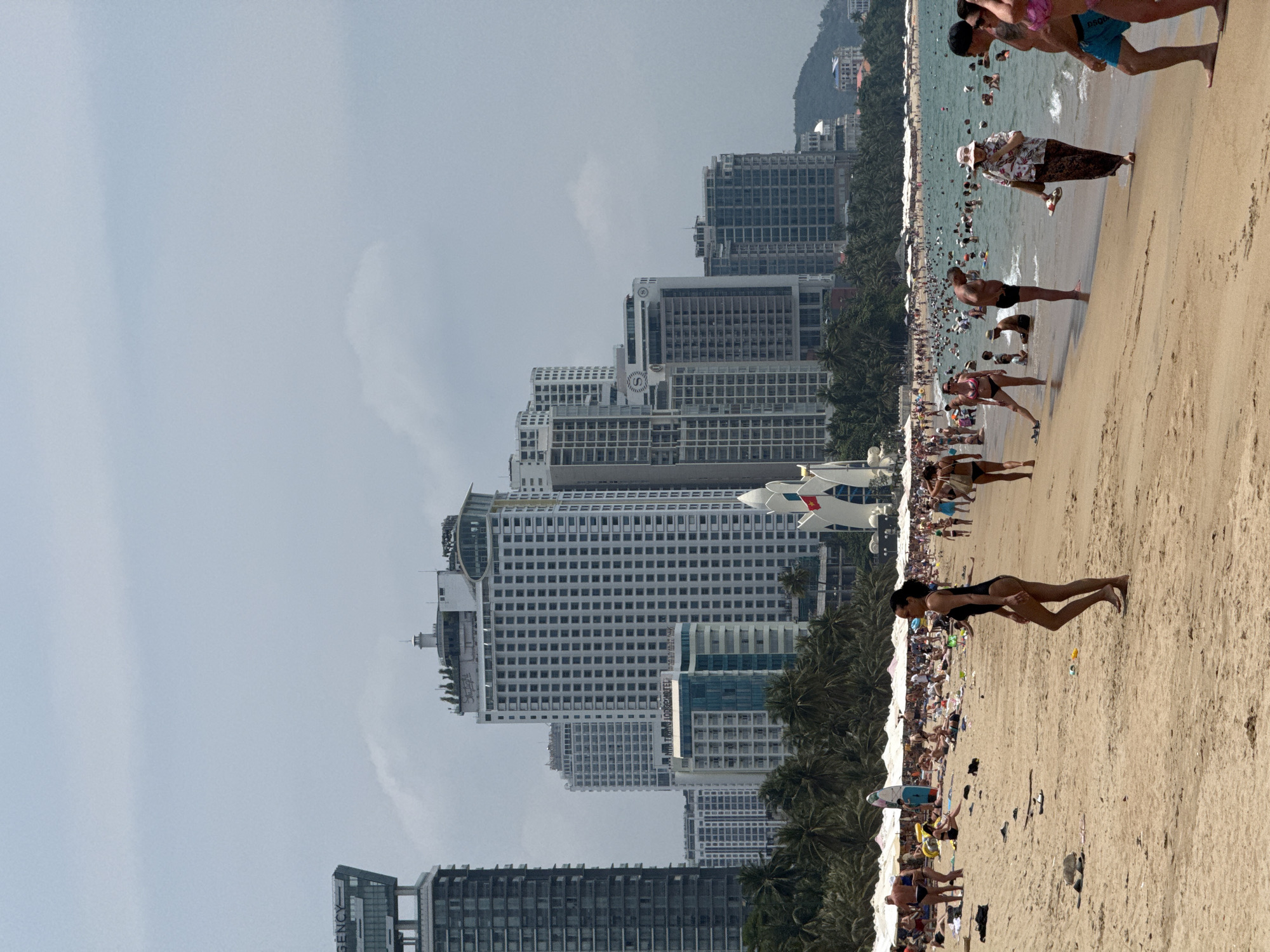 The image depicts a vibrant, bustling urban beach scene, likely taken on a sunny day. The perspective places the viewer looking across the sandy beach towards the ocean, with a dense city skyline lining the coast in the background.
Numerous people are visible across the frame, engaged in typical beach activities. Many are relaxing on the golden sand, some under beach umbrellas (partially visible in the distance), while others are walking or standing near the water's edge. A large number of individuals are seen wading or swimming in the blue-green ocean, which shows gentle waves. People are dressed in swimsuits and casual beach attire, indicating a leisure or holiday atmosphere.
The background is dominated by a long stretch of high-rise buildings, many appearing to be hotels or apartment blocks, interspersed with lush green palm trees. The architectural styles vary, with some modern glass facades and other more traditional multi-story structures. A mountainous or hilly landscape can be seen faintly in the distant background beyond the city.
The sky is bright, suggesting daytime, possibly mid-morning or afternoon, and appears mostly clear, though with a slight haze above the city. The overall mood is one of relaxation and enjoyment.
Several pieces of text are visible on the buildings:
- An "S" logo, possibly indicating a hotel chain, is visible on one building.
- The word "REGENCY" is seen vertically on the side of another modern building.
- On a white high-rise building, a small flag resembling the Vietnamese flag (red with a yellow star) is flying.
- Below the flag on the same building, "DSQUARED2" is visible, likely an advertisement or branding on the facade.
