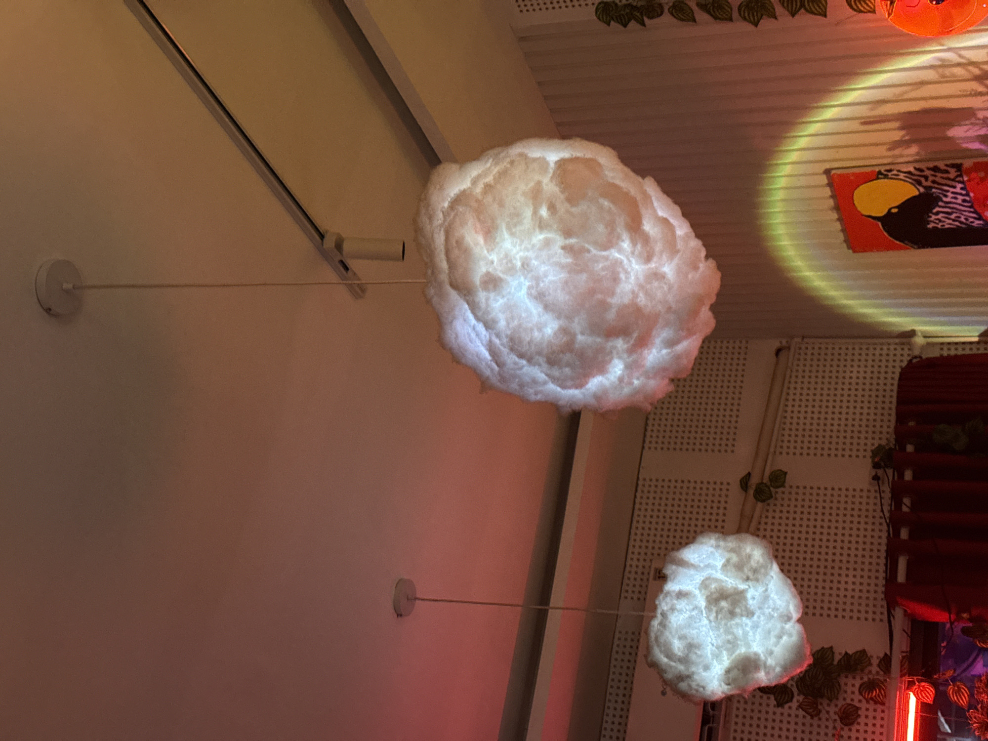 The image presents an interior view of a space with distinctive lighting fixtures shaped like clouds. The clouds are illuminated from within, providing a soft, diffused glow. One is large and dominates the upper portion of the frame, while a smaller cloud is visible in the lower right. These cloud fixtures are suspended from the ceiling and walls by thin rods connected to circular mounts.
The ceiling has a lattice-like structure and is decorated with artificial leaves, creating a whimsical ambiance. A painting of a bird with yellow and black markings on a red background can be seen on the ceiling. Further elements of the decor include a red wooden structure with vine-like embellishments and neon lighting.
The left side of the image is dominated by a plain wall with a track lighting fixture. The overall aesthetic suggests a modern, artistic space.
I am unable to provide the specific location in Đà Nẵng, Vietnam.