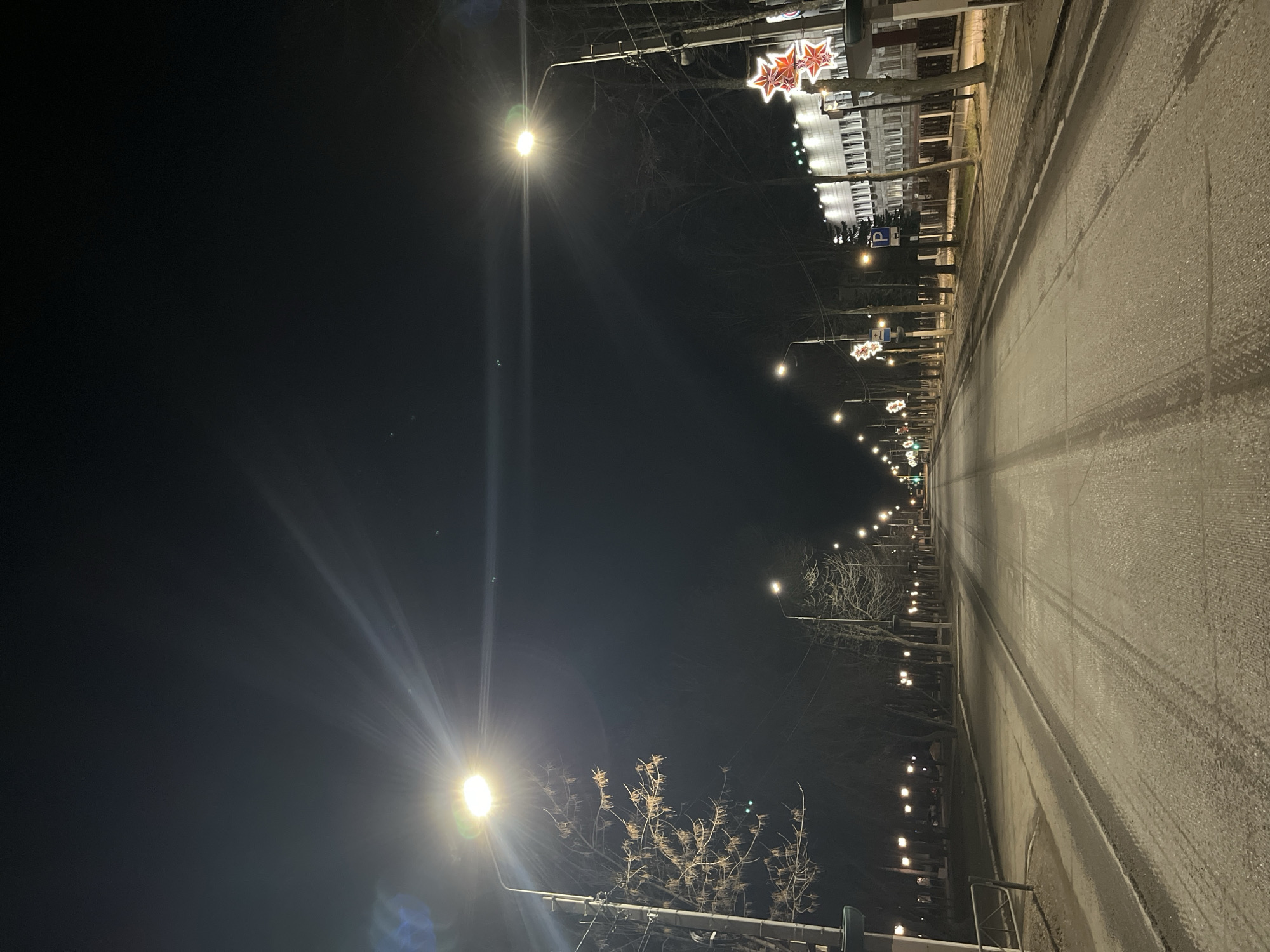 The image depicts a street scene at night in Chaikovsky, Russia. The street is illuminated by streetlights lining both sides, spaced evenly apart. Some of the lights feature red star-shaped decorations. The street itself appears wet or icy, reflecting the light. Tire tracks are visible on the right side of the road. On the right side of the street is a multi-story building. The sky is dark and clear, with no visible stars or clouds. The overall atmosphere is quiet and deserted.