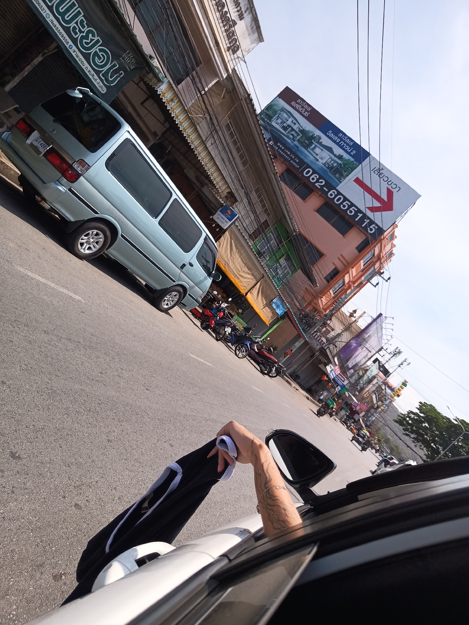 The image, slightly angled, depicts a street scene in Hua Hin City Municipality, Thailand, as seen from the passenger side of a car. A light blue Toyota van is parked on the left side of the road, next to a storefront with Thai lettering. Several motorcycles are parked behind the van. A large billboard with a red arrow indicating a right turn and the phone number 062-6055115 is prominently displayed on a building. Overhead power lines are visible. An arm extends from the car window, holding a black and white garment. The rearview mirror of the car is also visible.