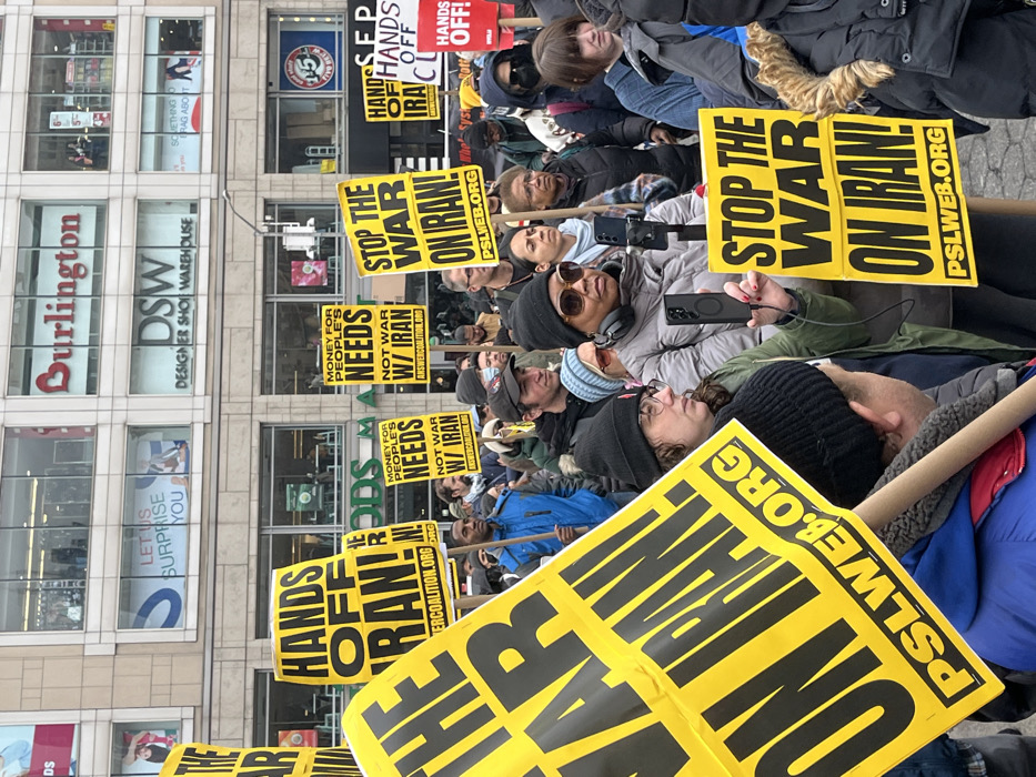 This image captures a public demonstration taking place in an urban commercial district of New York City, United States of America. A large gathering of people, predominantly adults, are visible, dressed in various winter garments including jackets, beanies, and scarves, indicating cold weather. The individuals are holding numerous bright yellow protest signs with black block lettering. Prominent messages displayed on these signs include "STOP THE WAR ON IRAN!", "HANDS OFF IRAN!", and "MONEY FOR PEOPLE'S NEEDS NOT WAR W/ IRAN." Many signs also feature web addresses such as "PSLWEB.ORG" and "ANSWERCOALITION.ORG."
In the foreground, a woman wearing a grey puffer jacket, dark sunglasses, and headphones holds a black smartphone vertically in her right hand, extended forward as if recording or photographing the event. A cable connects from the phone to a small device mounted on a stick she also holds. To her immediate left, another woman with glasses and a black beanie is partially visible, holding a large yellow sign. The crowd appears diverse and is generally facing forward.
The background features multi-story commercial buildings. Clearly visible storefronts include "Burlington" on an upper floor and "DSW Designer Shoe Warehouse" below it. Below DSW, part of a sign displaying "GOODS" is visible, likely indicating another retail store. A window sign on an upper floor reads "LET US SURPRISE YOU." The sky above is overcast.