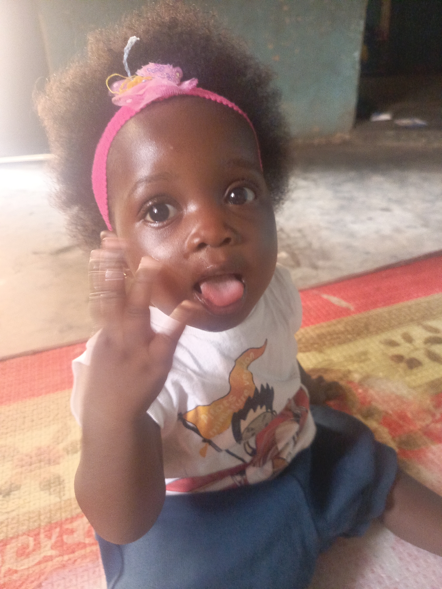 A low-angle, close-up shot depicts a young child sitting on a patterned surface. The child, with dark skin and curly hair adorned with a pink headband, sticks out their tongue while holding up their left hand with fingers spread apart. The child wears a white t-shirt with a cartoon graphic. The location appears to be indoors based on the wall behind the child, potentially a residential setting within Akwanga, Nigeria. The patterned surface beneath the child appears to be a rug.