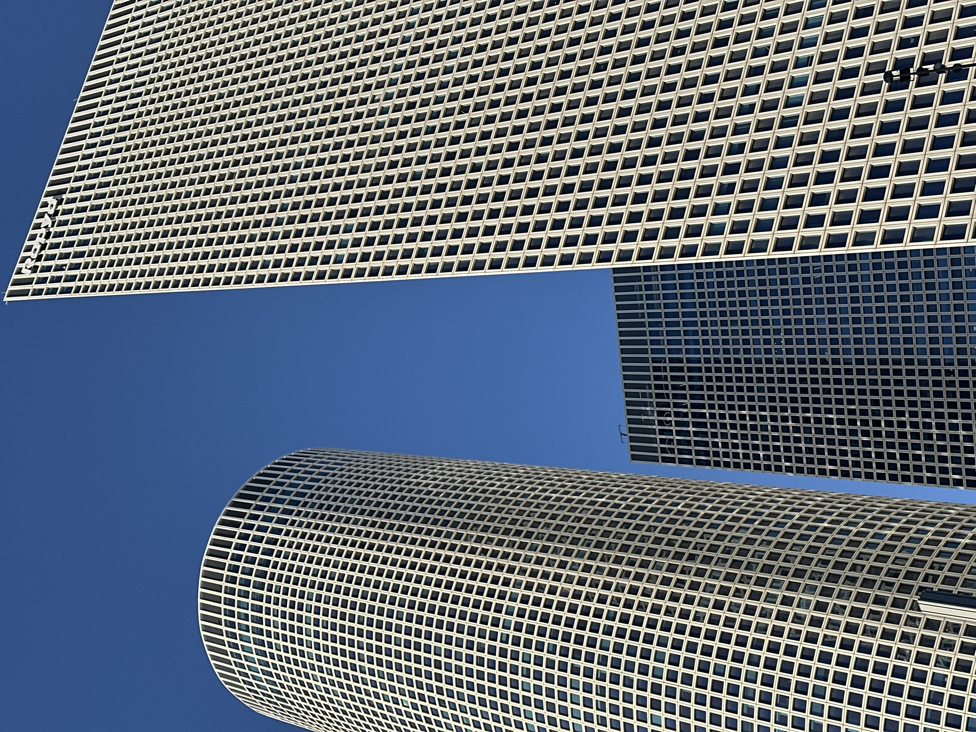 The image presents an abstract architectural composition of three modern high-rise buildings set against a vivid, cloudless blue sky, characteristic of Tel Aviv, Israel. The photograph is taken from an upward perspective, highlighting the geometric patterns and contrasting shapes of the structures.
In the upper-left quadrant, a prominent rectangular building features a grid-like facade composed of numerous square windows within a light, possibly off-white, structural framework. Along its visible left edge, the word "AZRIELI" is clearly discernible, spelled out vertically in white letters.
Towards the bottom right, a large cylindrical building occupies a significant portion of the frame, showcasing a similar intricate grid pattern of square windows and light framework that curves with its shape.
A third rectangular building is visible on the middle-right side, positioned between the other two. This structure appears darker, likely due to its reflective glass facade, but also displays a consistent grid of windows.
The sky is a clear, deep blue, suggesting excellent weather conditions. The strong, direct lighting indicates a sunny mid-day, casting minimal shadows and enhancing the crispness of the architectural lines. No people or specific activities are visible, allowing the focus to remain on the striking interplay of modern design and the expansive sky.