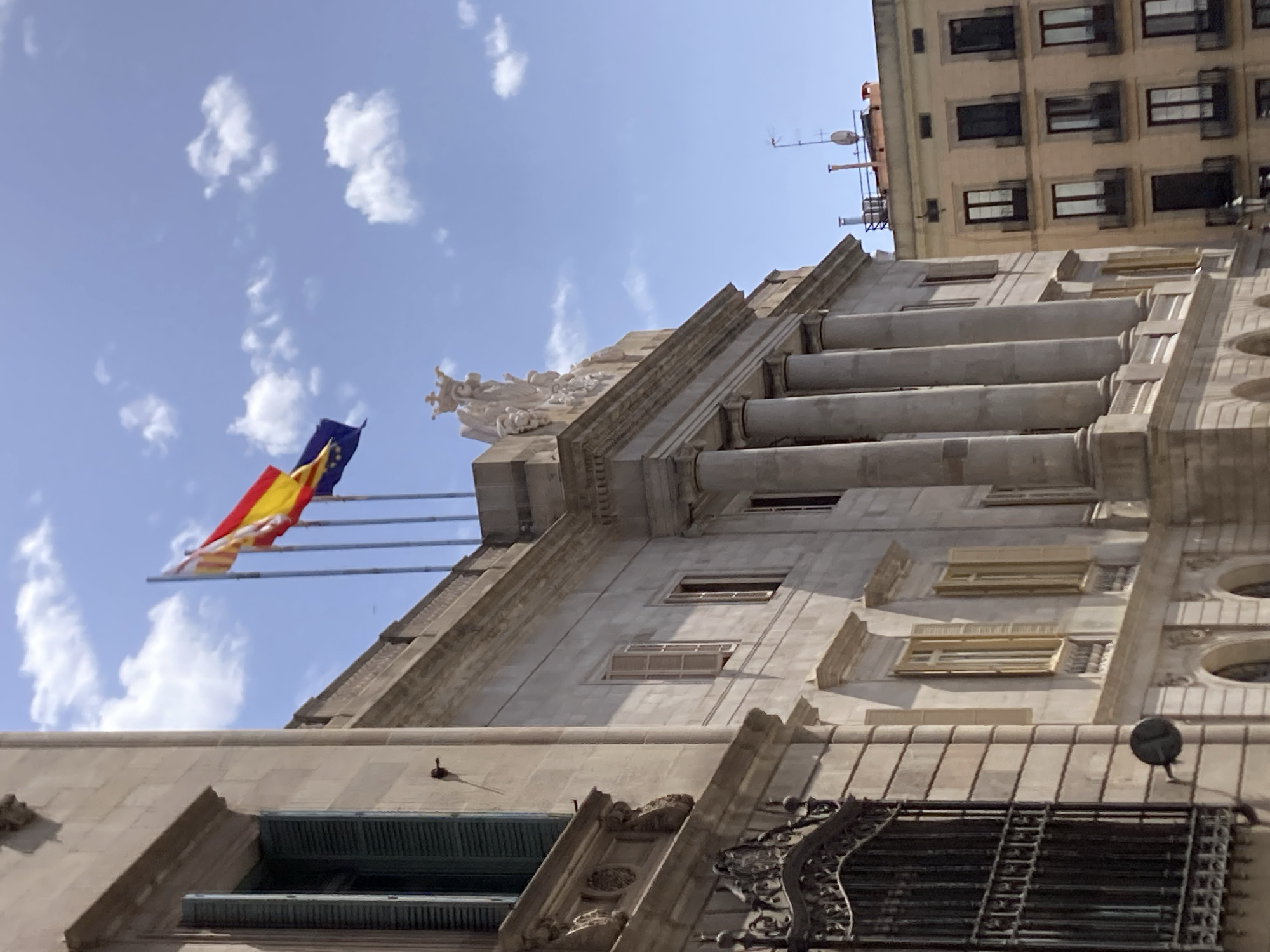 The image captures a section of the Palau de la Generalitat de Catalunya, located in Barcelona, Spain. The building's facade is constructed from light-colored stone, featuring large columns and ornate architectural details. Several windows with open shutters are visible. Three flags are flying on flagpoles atop the building: the flag of Catalonia, the flag of Spain, and the flag of the European Union. The sky above is blue with scattered white clouds. To the right, a more modern, yellow-colored building with multiple windows and balconies stands adjacent to the historic structure. The overall scene depicts a sunny day in a historic European city, highlighting the juxtaposition of old and new architecture.