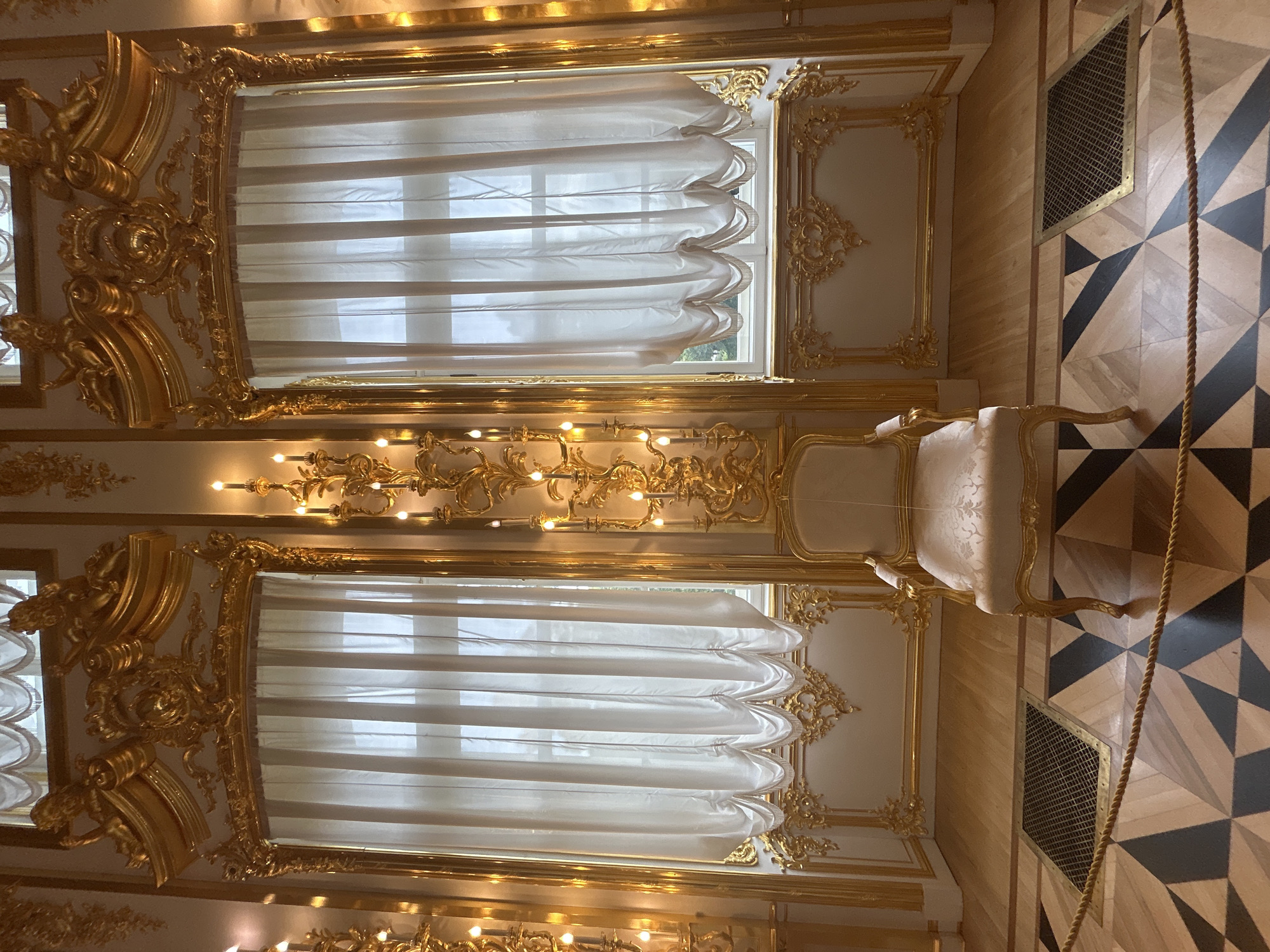 This is a medium shot, eye-level, indoor photograph of a richly decorated room. The scene is dominated by ornate gold trim and elaborate wall sconces. Two large windows with sheer white curtains hang in the background, framing what appears to be daylight outside. In the center foreground, a single, cream-colored armchair with gold trim sits on a polished wooden floor. The floor features a geometric pattern of light wood and dark diamond shapes. A metal grate is visible on the floor to the left of the chair. A thick, golden rope barrier runs across the foreground of the image. The room appears to be a museum or a historical palace, possibly located in a city like St. Petersburg, Russia, given the opulent style often associated with the Winter Palace. The lighting is soft and diffused, likely from the windows and the illuminated sconces. There are no people visible in the image.