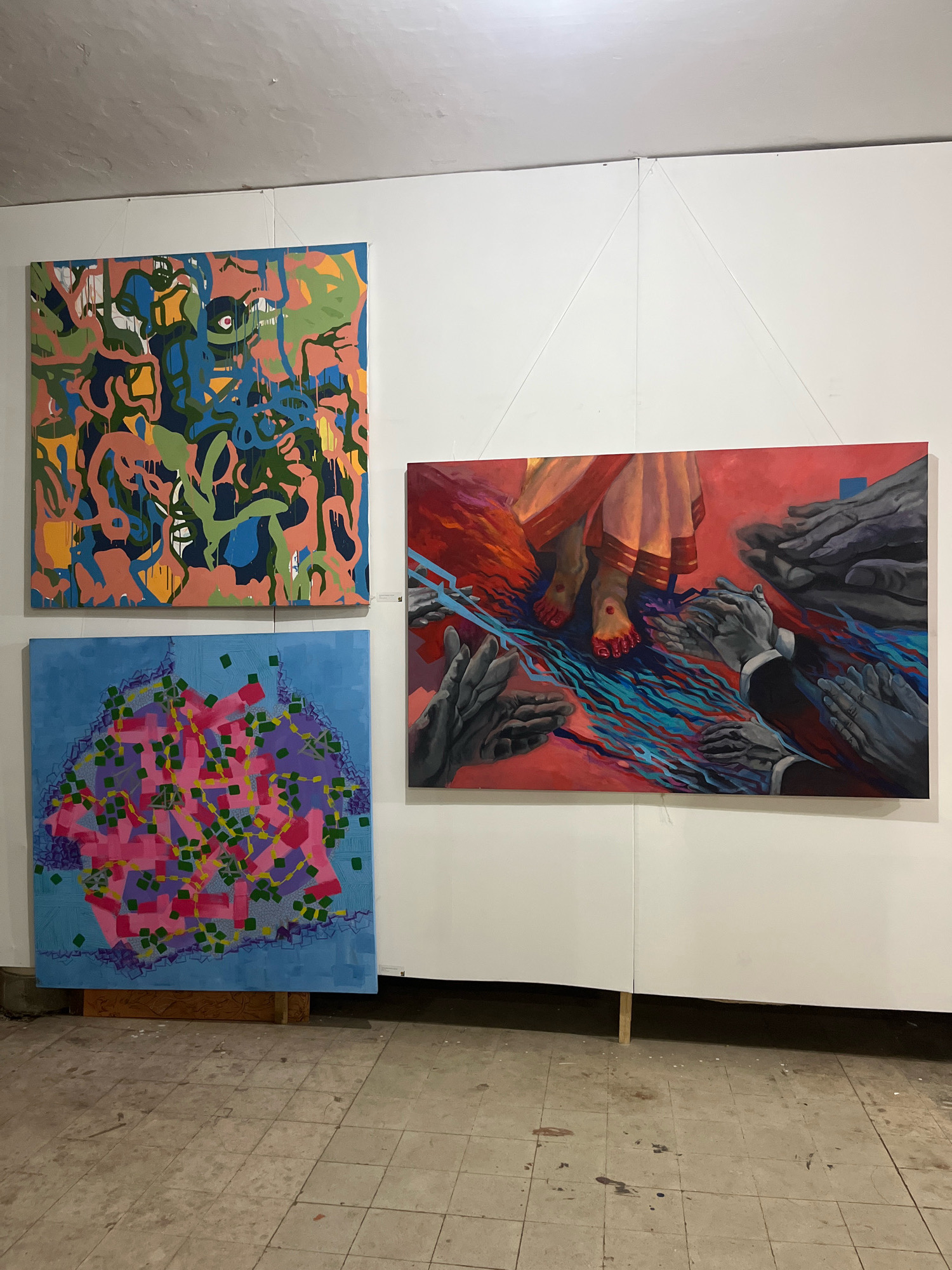 This image depicts an art exhibition in Dhaka, Bangladesh. Three paintings are displayed on a white wall. The painting on the top left is abstract, with a chaotic mix of colorful organic shapes in blue, green, pink, and yellow. The painting on the bottom left is also abstract, featuring a blue background with a cluster of pink and purple geometric shapes overlaid with green squares. The painting on the right is a figurative work depicting a pair of feet in traditional Indian attire standing on a red, wavy surface. Surrounding the feet are several pairs of hands rendered in grayscale, reaching towards the viewer. The floor is tiled and appears worn, with some paint splatters. The overall environment suggests an indoor exhibition space with simple wall mounting.