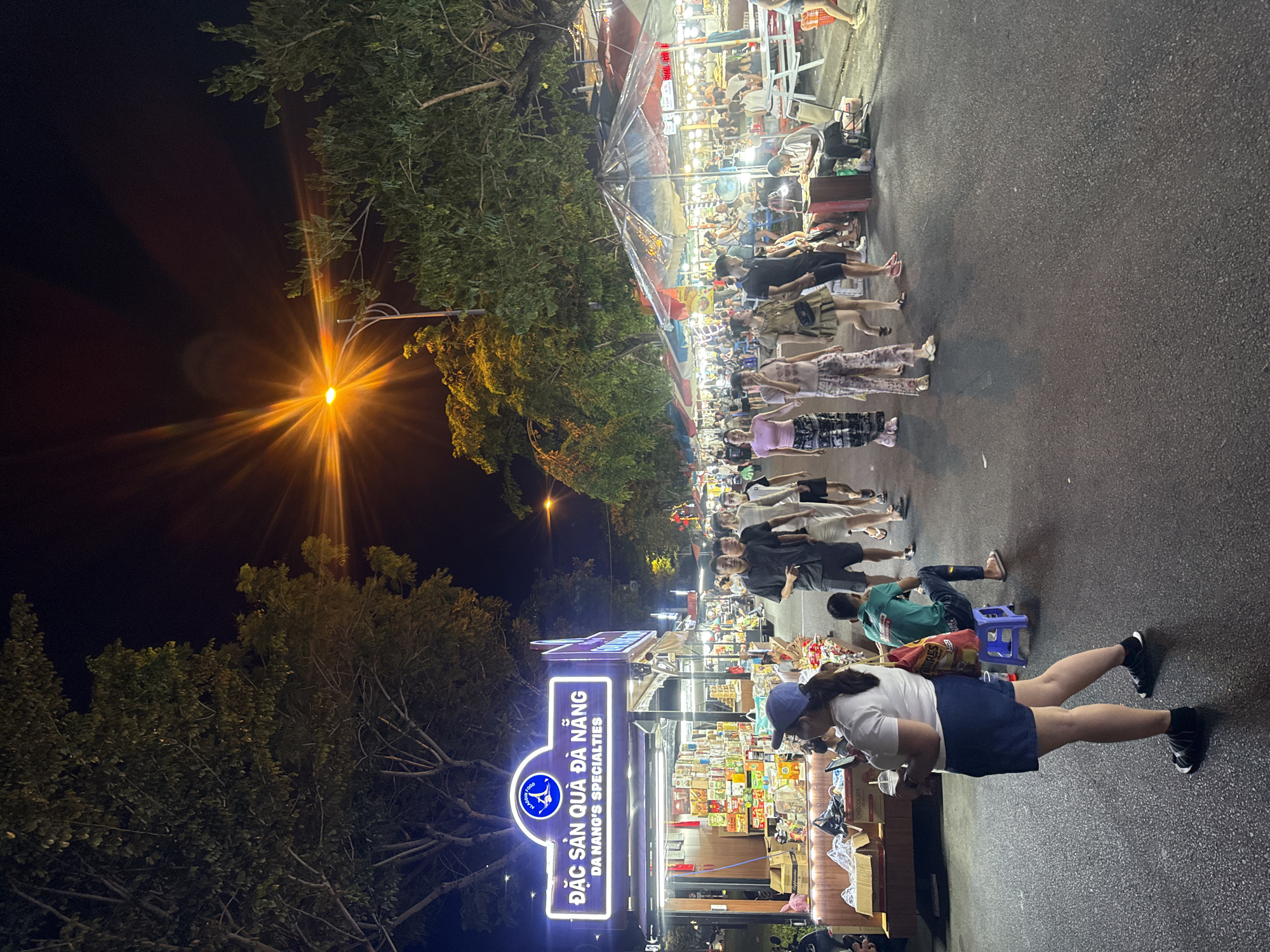 The image shows a night street scene with pedestrians walking and vendors selling goods. A bright street light is visible on the left of the image, illuminating trees. A store sign reading "Đặc Sản Quà Đà Nẵng" is also visible. In the foreground, a person is standing near a vendor's stall. Other people are visible along the street, browsing and walking. The scene takes place in Đà Nẵng.