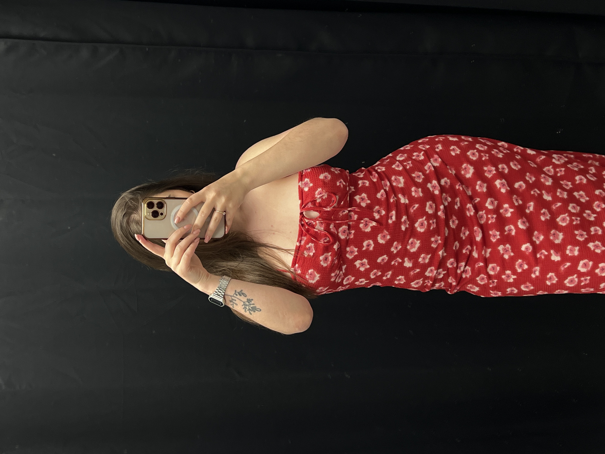 A woman is taking a selfie. She is wearing a red dress with a white floral pattern. She is holding her phone in her left hand and the phone covers her face. She is wearing a ring on her left ring finger. She is wearing a watch on her left wrist and a floral tattoo below the watch. The background is black and appears to be a wall. There is no evidence to suggest this image was taken in Ternopil, Ukraine.