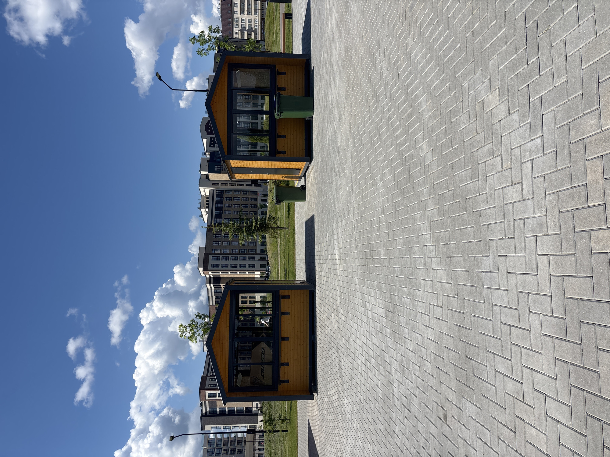 A view of an outdoor area in Varaksino, Russia, shows two small wooden structures, possibly shelters or kiosks, positioned on a paved pathway made of gray bricks laid in a herringbone pattern. These structures have black frames, clear glass panels, and peaked roofs. A green trash can is visible inside one of them. In the background, modern apartment buildings rise several stories high. A blue sky with scattered puffy white clouds is visible above the buildings. A lamppost stands near the edge of the pathway. The image is oriented with the sky on the left and the pavement on the right.