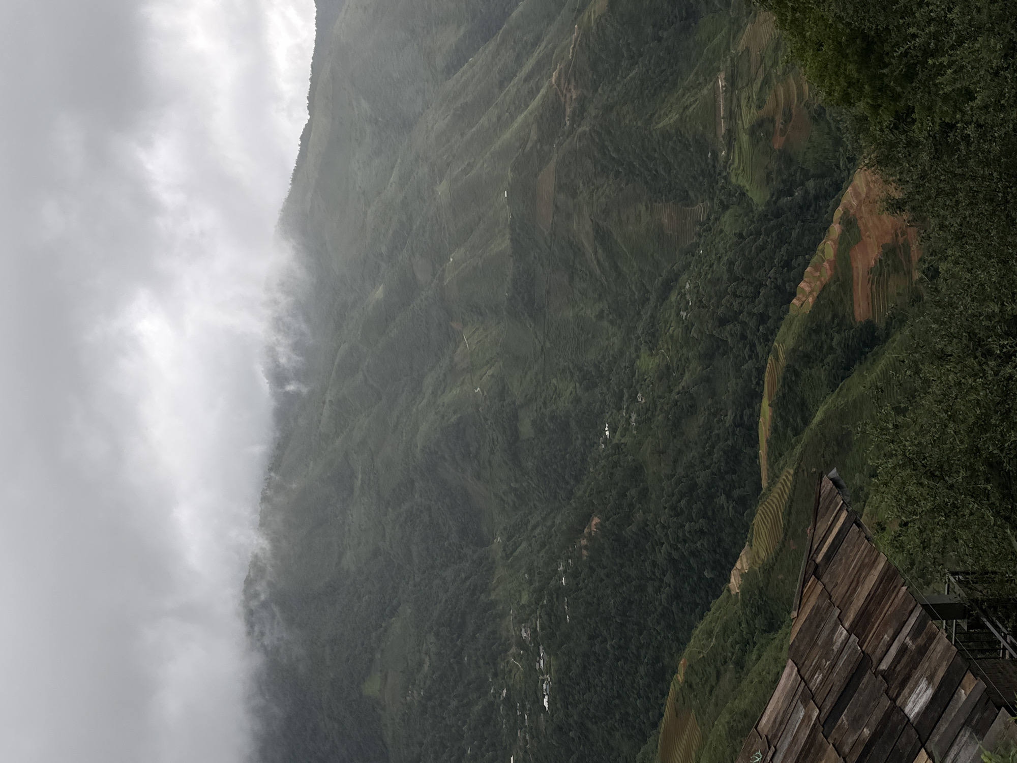 The image captures a sweeping vista of lush green mountains in Ta Xua, Vietnam, under a heavy, overcast sky. The mountains are characterized by their steep slopes, extensively terraced for cultivation, revealing patches of tilled brown earth interspersed with verdant foliage. Scattered along the hillsides are small clusters of buildings, suggesting human settlement amidst the natural landscape. In the foreground, a portion of a rustic wooden roof is visible, hinting at a structure from which the photograph was taken. The overall atmosphere is one of serene, yet somewhat somber, natural beauty, typical of a cloudy mountain region.
