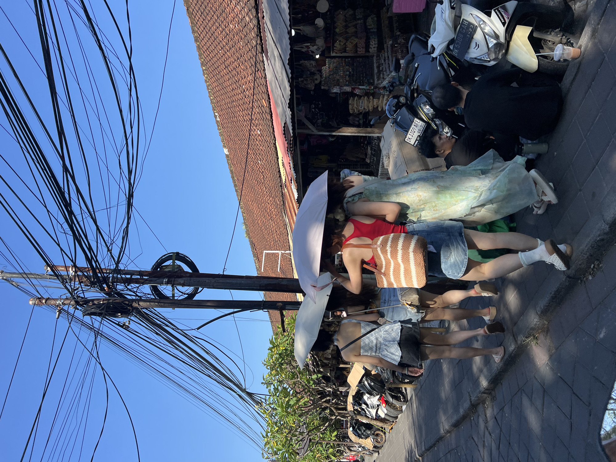 In Kuta, Indonesia, a street scene unfolds under a clear blue sky, dominated by a dense network of overhead electrical wires. Four individuals, three women and one person whose gender is not discernible, walk away from the camera. The women are holding up umbrellas, suggesting it is sunny and they are seeking shade. Two of the women are wearing denim skirts, one with a red top and a woven bag, the other in a patterned top. The third woman is wearing a light blue top and denim shorts. A fourth person, wearing a green dress, is partially obscured by the other individuals.
To the right of the walkers, a row of parked motorcycles is visible. Two men are seated on the curb next to a white Honda motorcycle, one appearing to be looking at the other. A cup with a straw sits near the motorcycle.
Behind the people and motorcycles, there is a row of shops with tiled roofs and awnings, displaying various goods. To the left of the scene, more parked motorcycles and trees line the sidewalk, which is paved with dark rectangular stones. The overall impression is a busy, outdoor market or shopping area in a tropical climate.