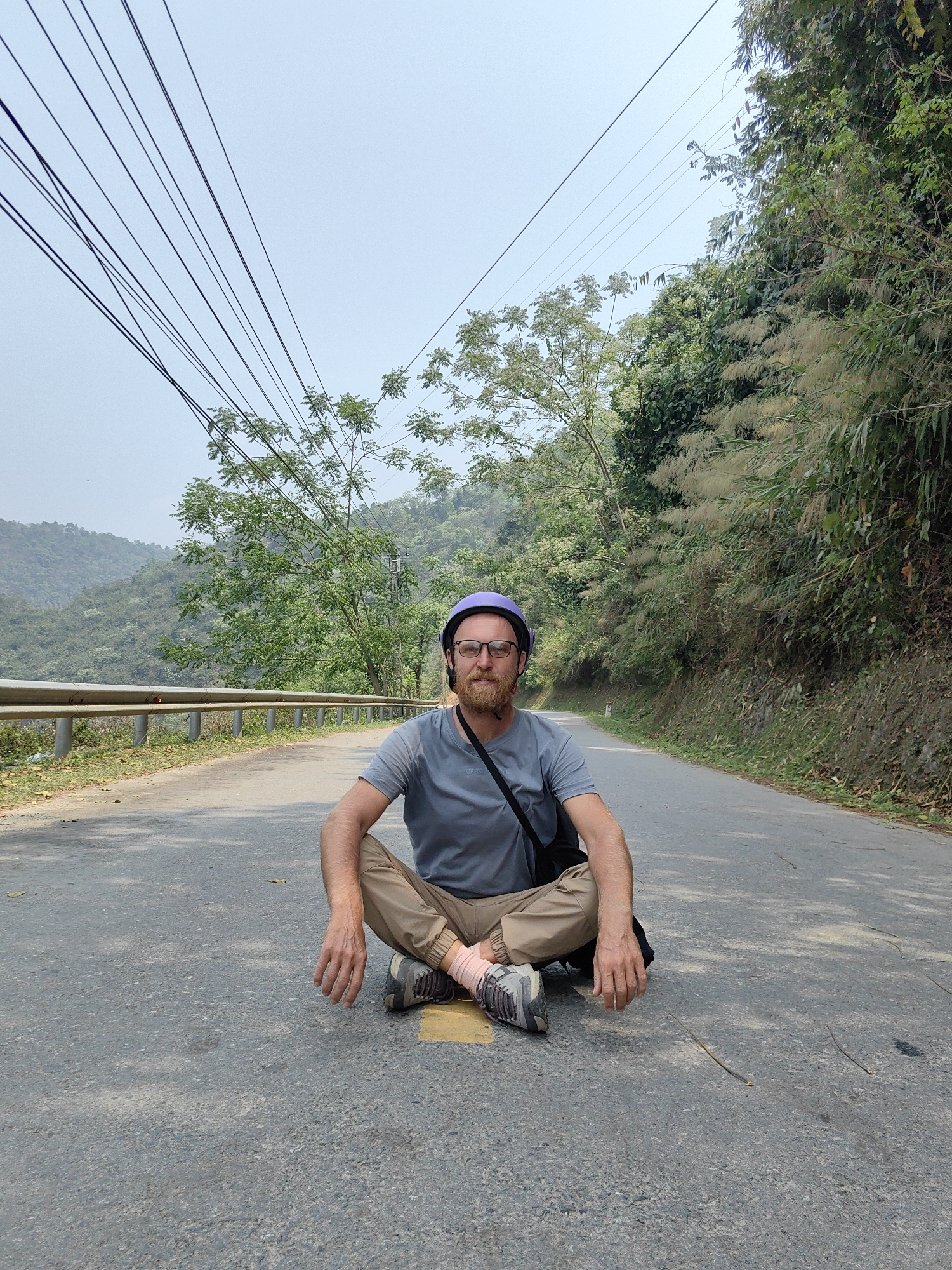 A man sits cross-legged on an asphalt road, facing the viewer. He wears a light purple helmet, glasses, a light grey t-shirt, and khaki pants. He has a beard and is wearing sneakers with pink socks. A black bag strap is across his shoulder. The road is flanked by vegetation, including trees and bushes. A metal guardrail is to the left. Power lines stretch across the sky above the road. The backdrop consists of green, mountainous terrain. The photo was taken on a road in Bảo Lạc, Vietnam.